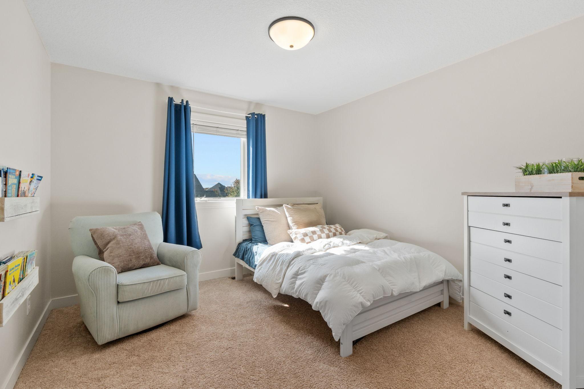 Bedroom with a white bed, blue curtains, beige carpet, and a cushioned armchair.