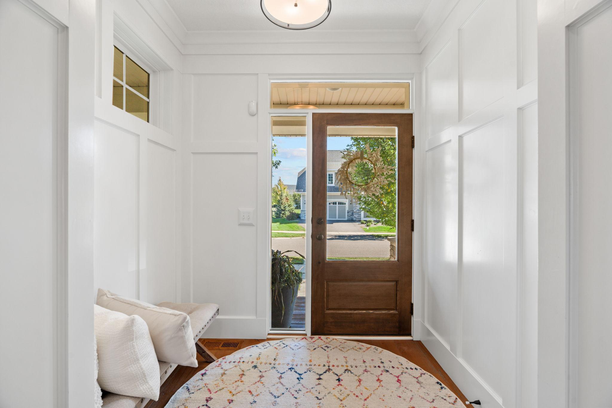 Entry foyer with a dark wood glass-paneled door, wreath, white paneling, and patterned rug.