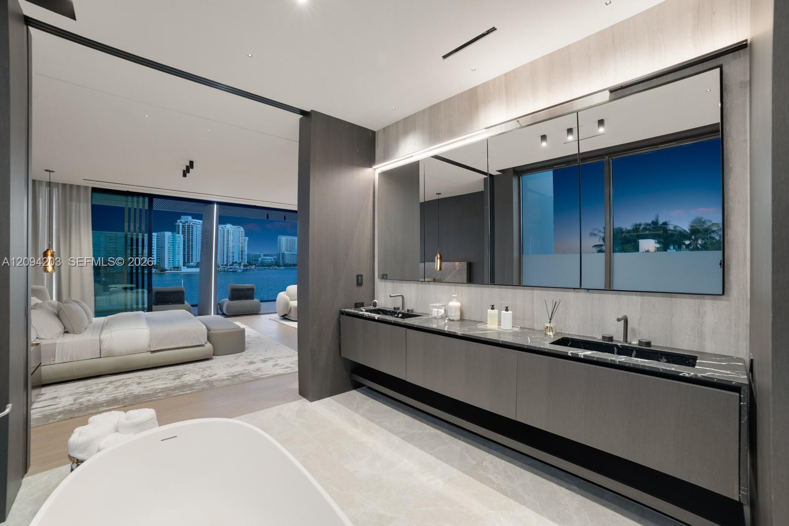 Modern ensuite bathroom with a freestanding tub and double vanity, opening to a bedroom overlooking the water.