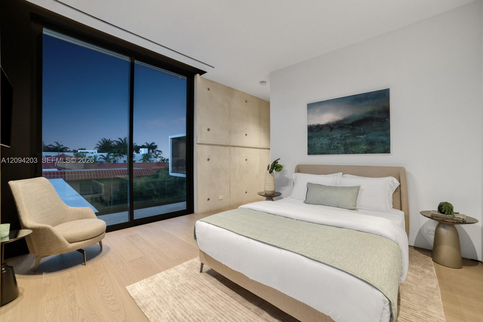 Modern bedroom with a large bed, concrete accent wall, and floor-to-ceiling windows overlooking palm trees.