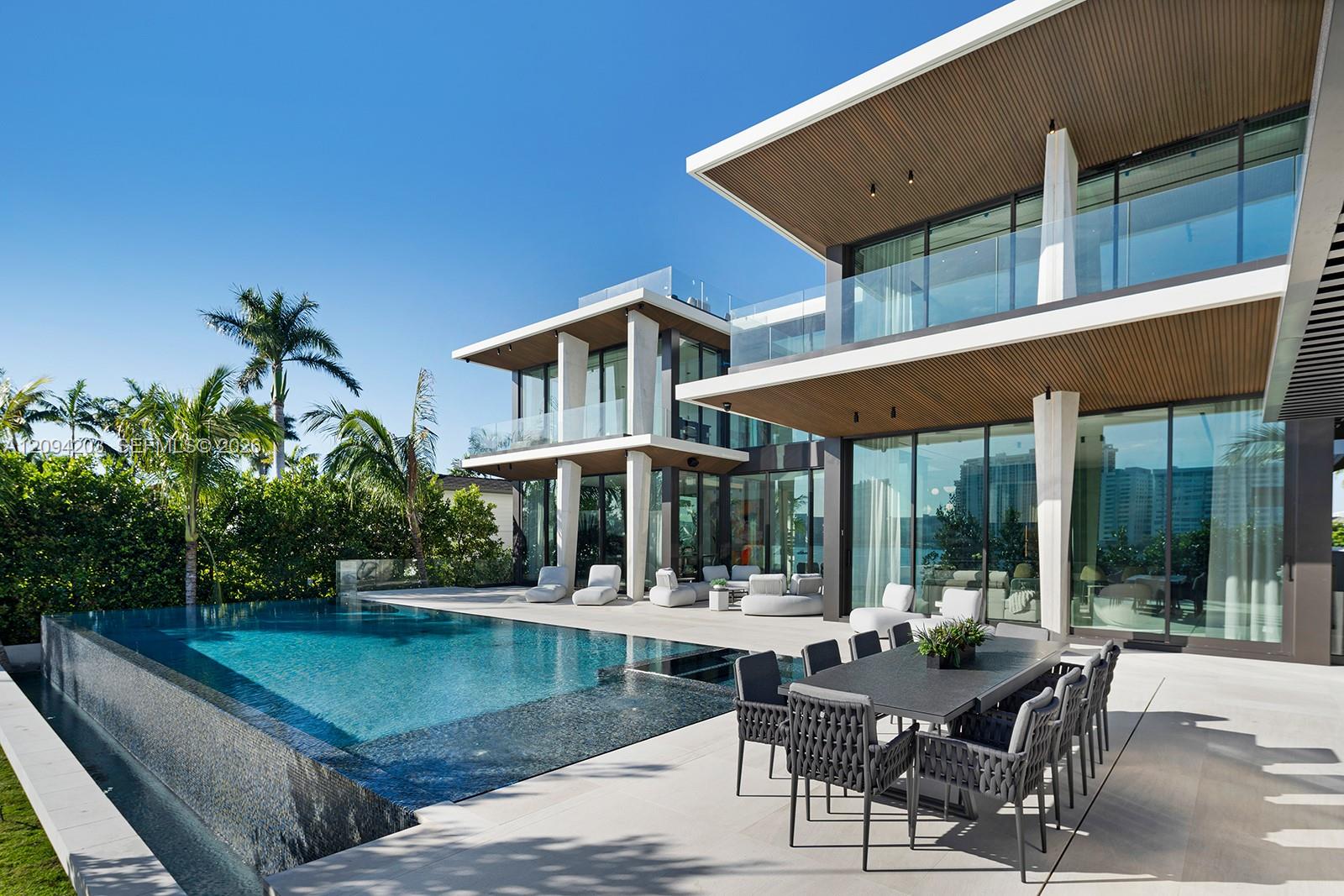 Modern glass-walled home beside an infinity pool, with palm trees and a furnished outdoor dining terrace.