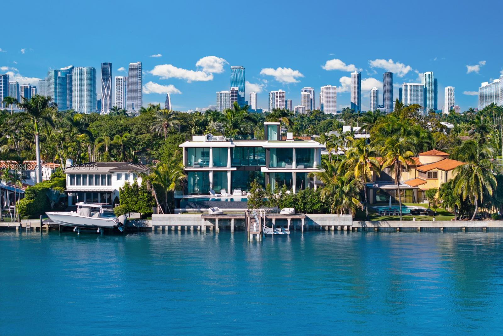Modern glass waterfront home with a dock and boat, palm trees, and a distant city skyline.