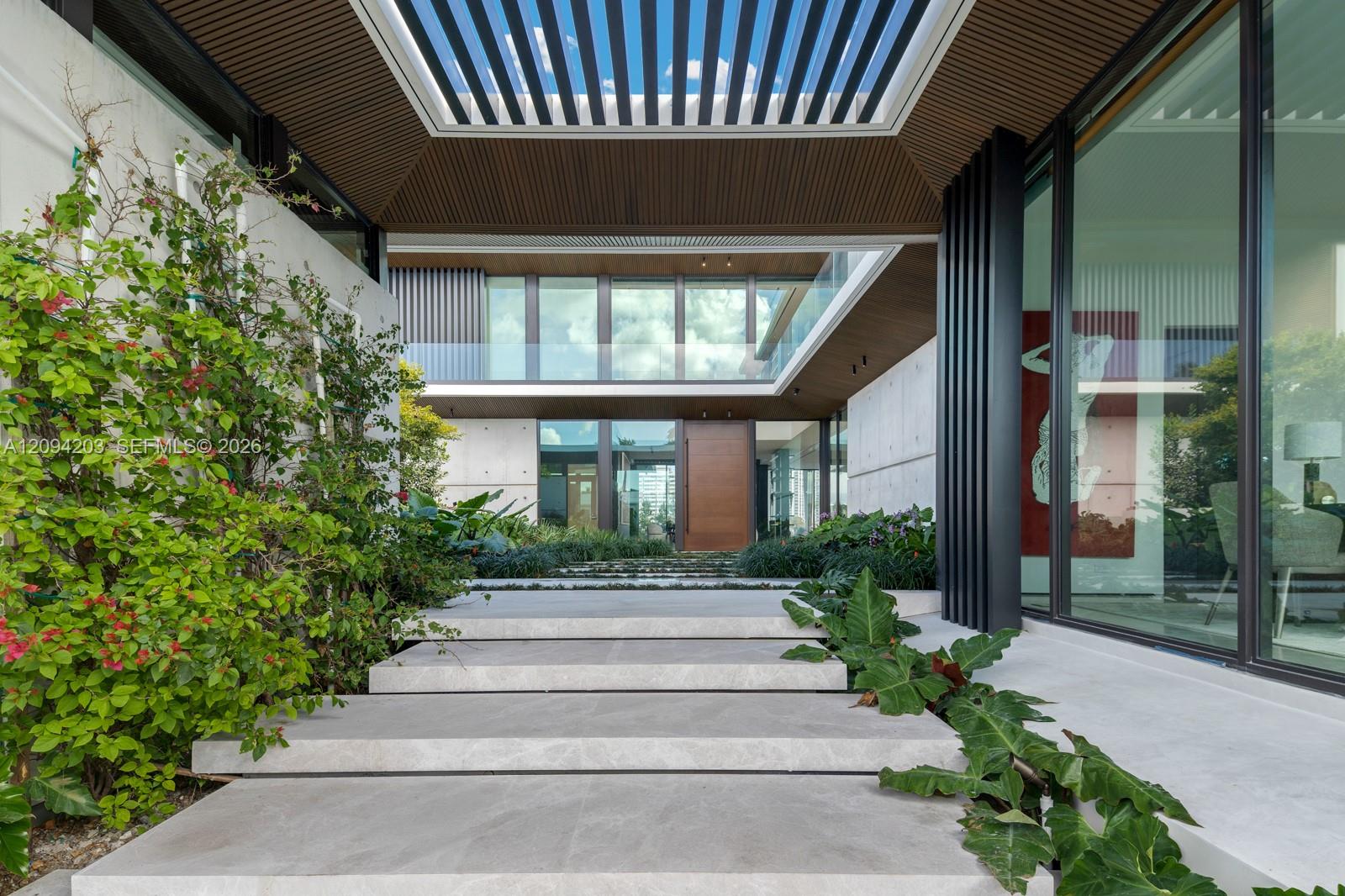 Modern home entrance with broad stone steps, tropical planting, glass walls, and a slatted wood canopy.
