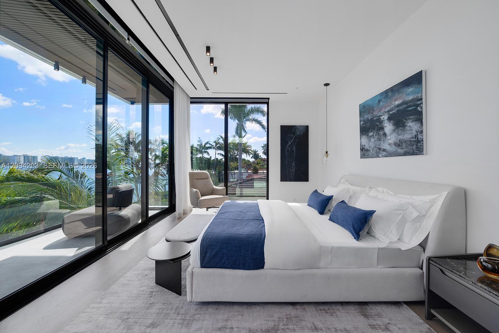 Contemporary bedroom with a white bed, blue accents, and floor-to-ceiling windows overlooking water and palms.