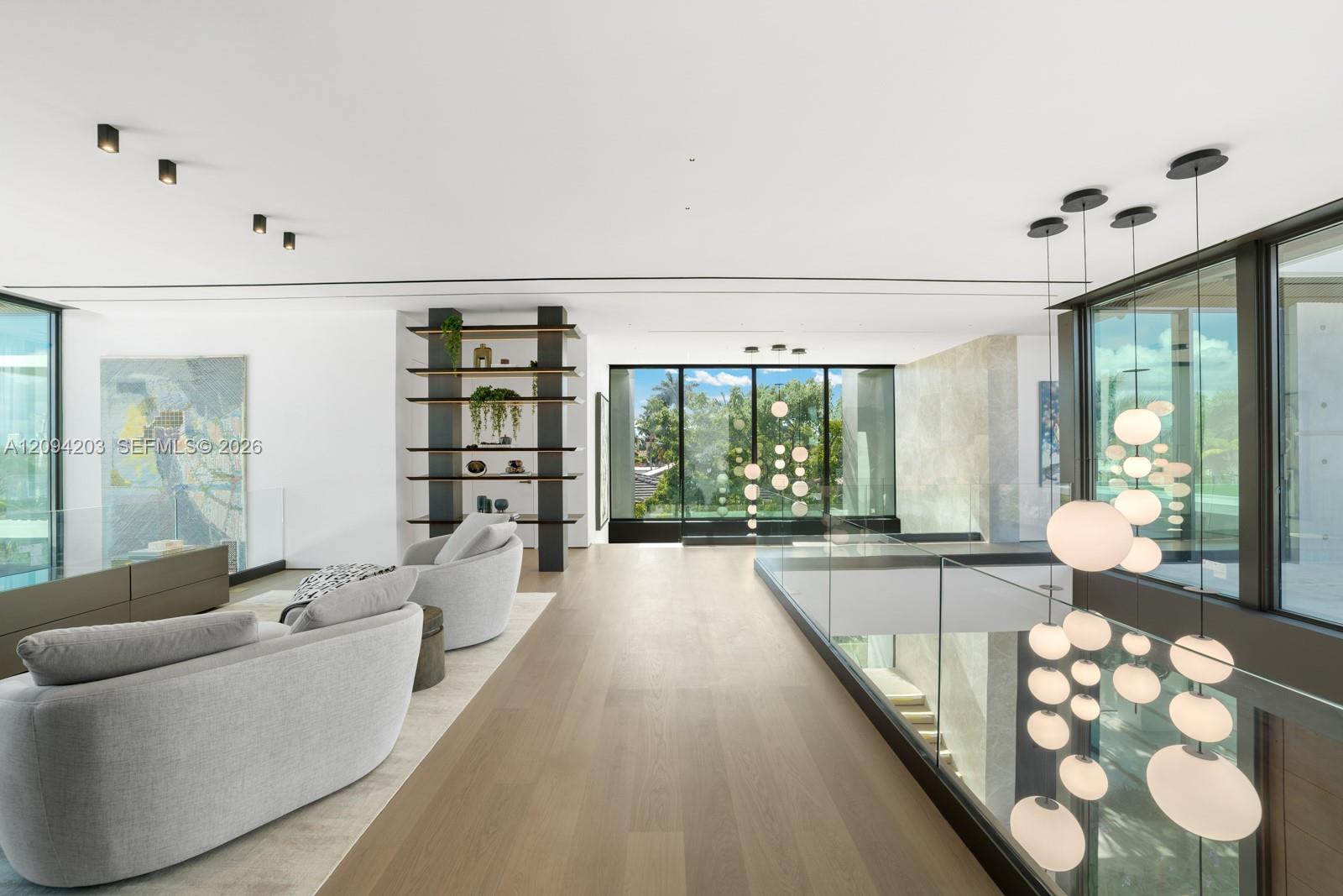 Modern living area with curved gray sofas, floor-to-ceiling windows, and a glass railing beside hanging globe lights.