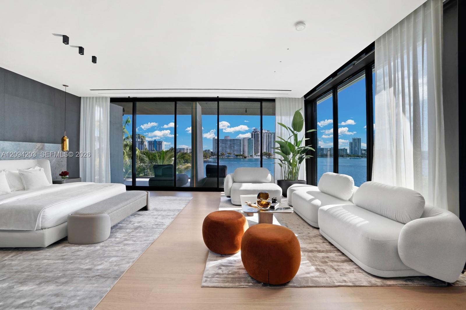 Modern bedroom with white lounge seating and floor-to-ceiling windows overlooking the water and skyline.