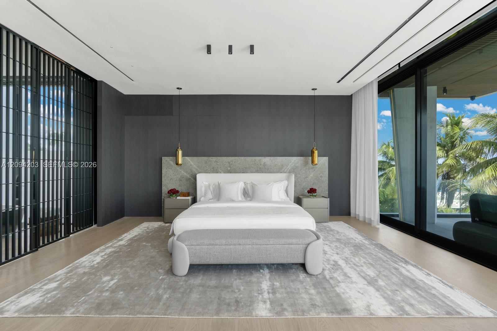Modern bedroom with a white bed, gray stone headboard wall, and glass doors overlooking palm trees.