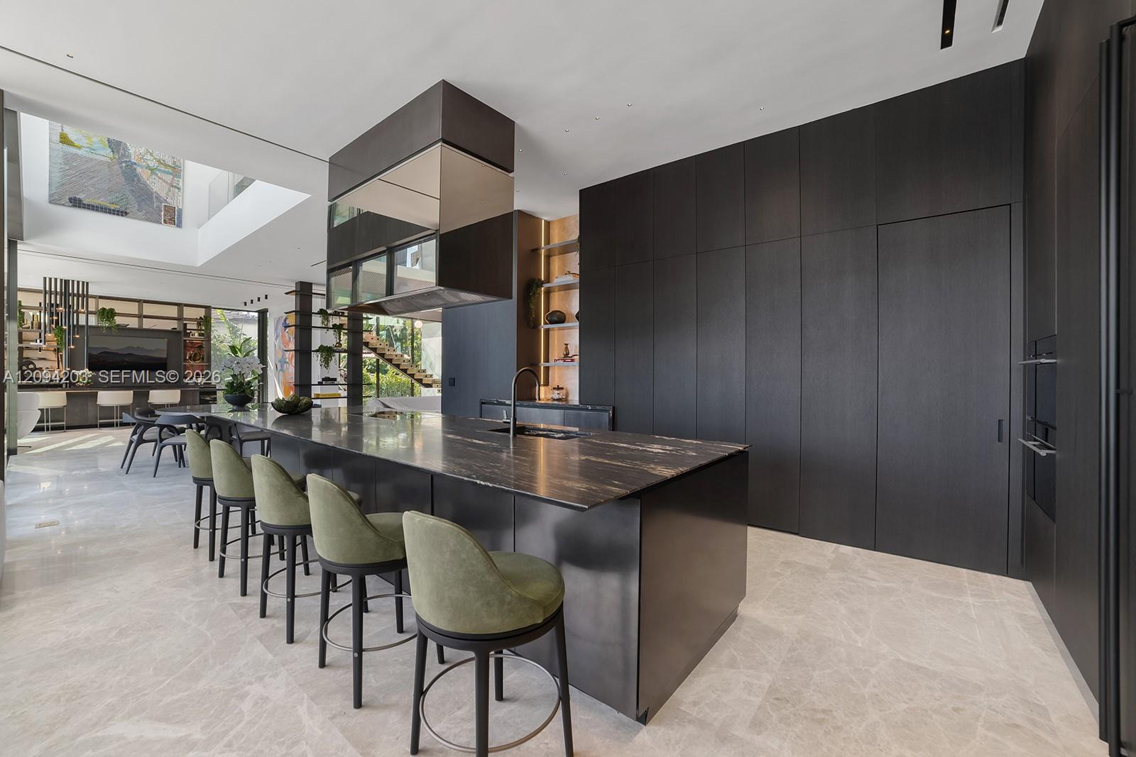 Contemporary kitchen with a large stone island, green bar stools, dark cabinetry, and a glass-sided staircase.