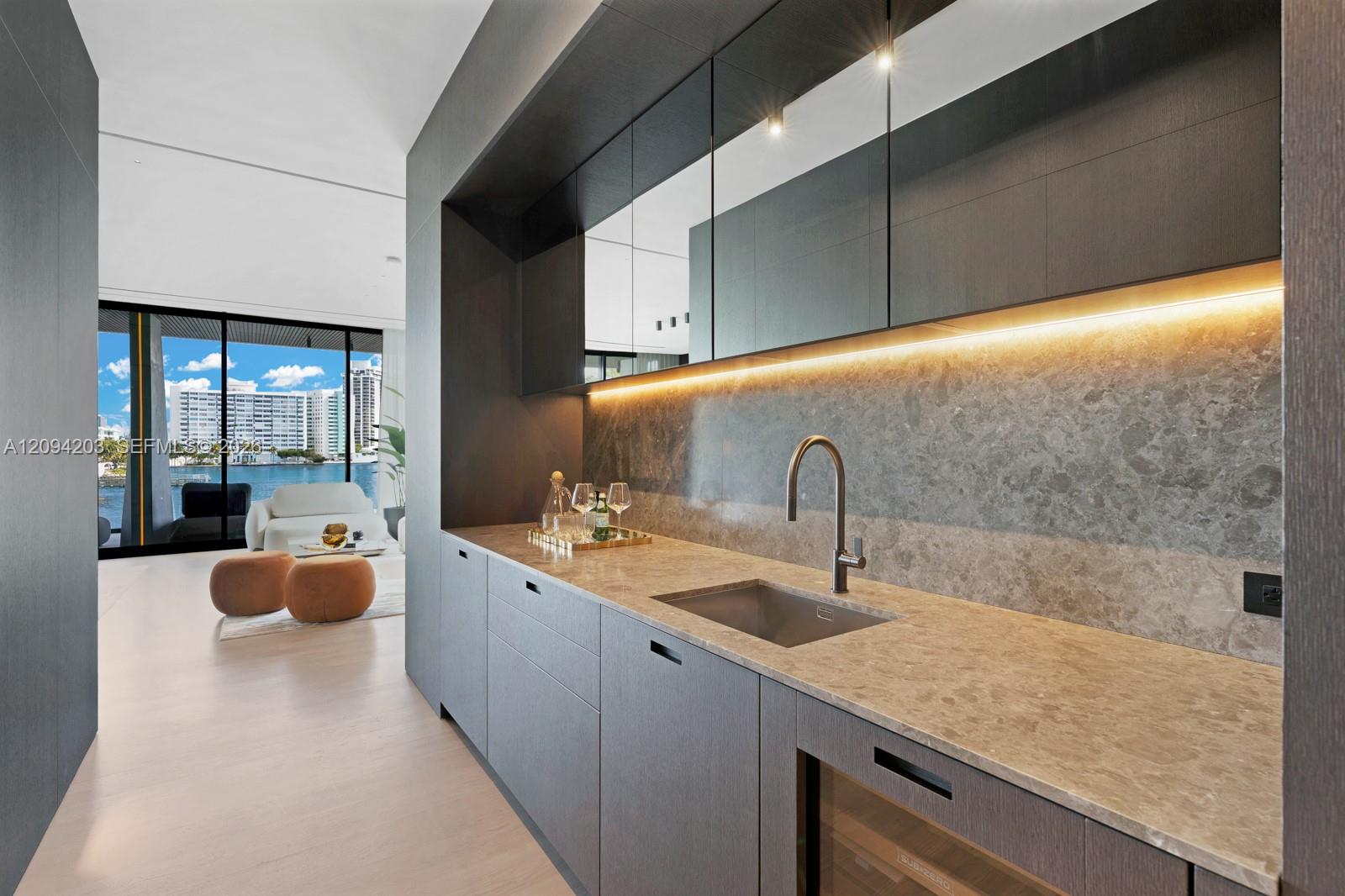 Modern kitchen with gray cabinetry and stone counters, opening to a waterfront living area.