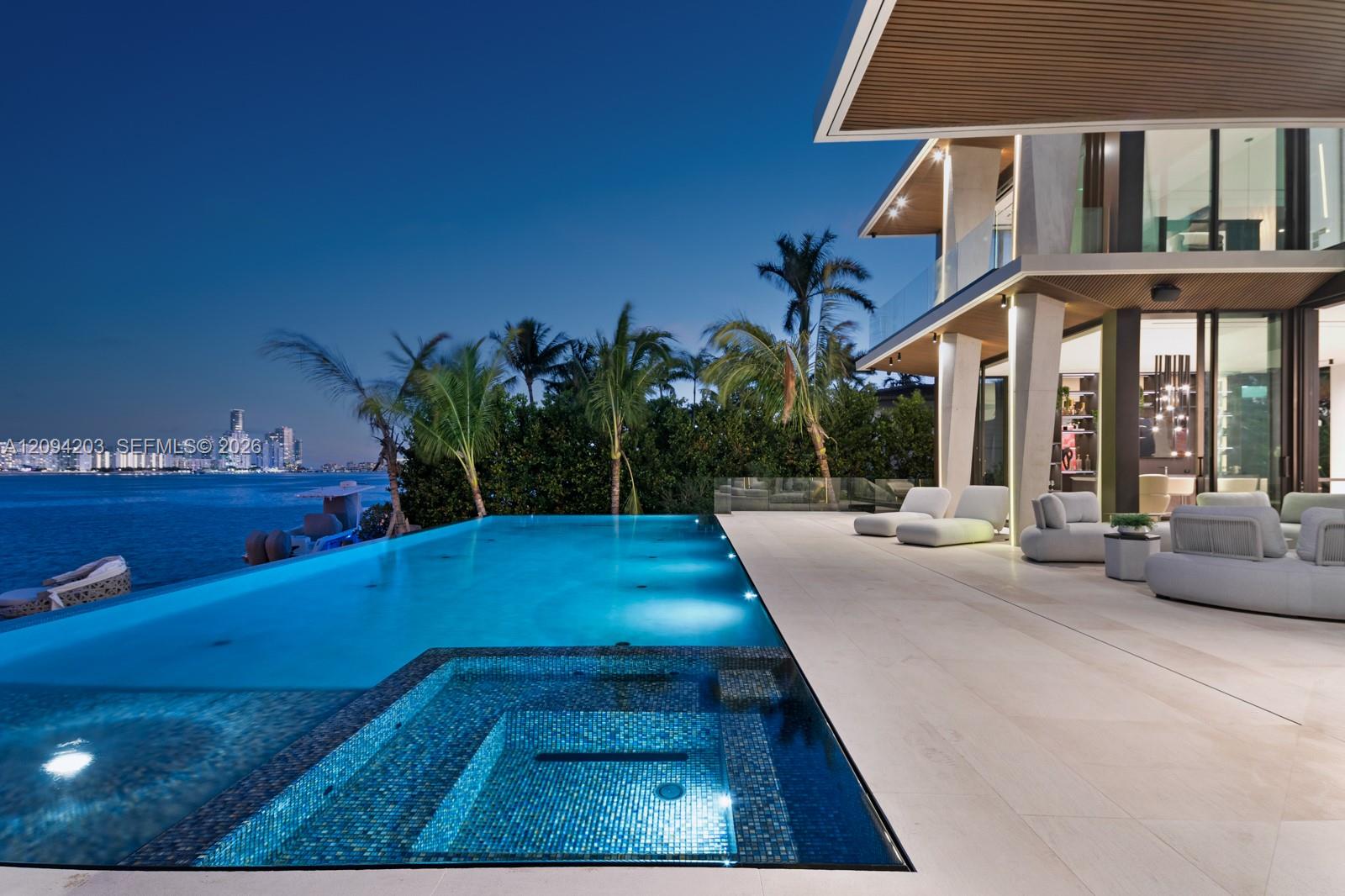 Waterfront home with a blue-lit infinity pool, patio seating, and city skyline at dusk.