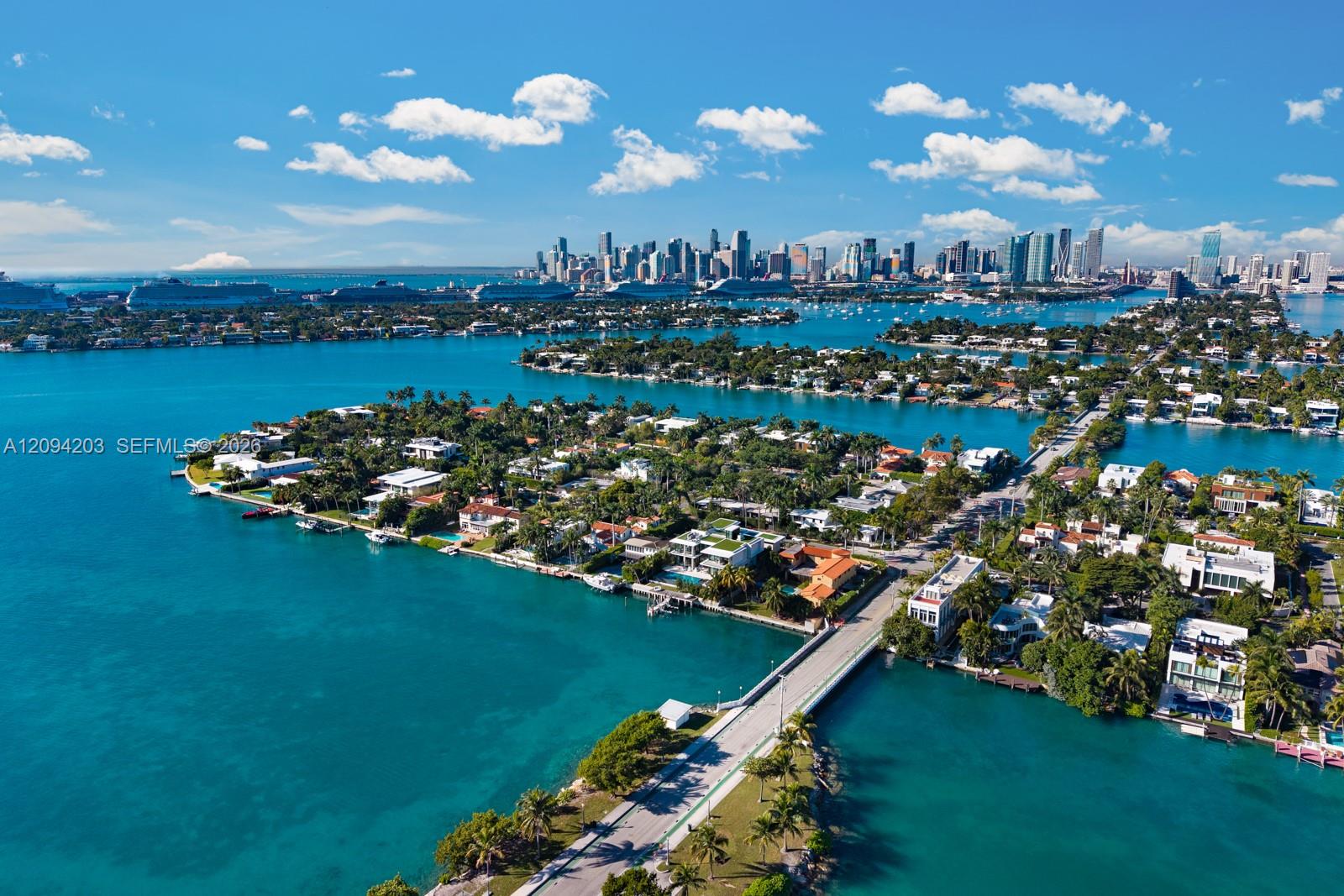 Aerial view of Miami Beach waterfront homes, turquoise waterways, a bridge, and the downtown skyline.