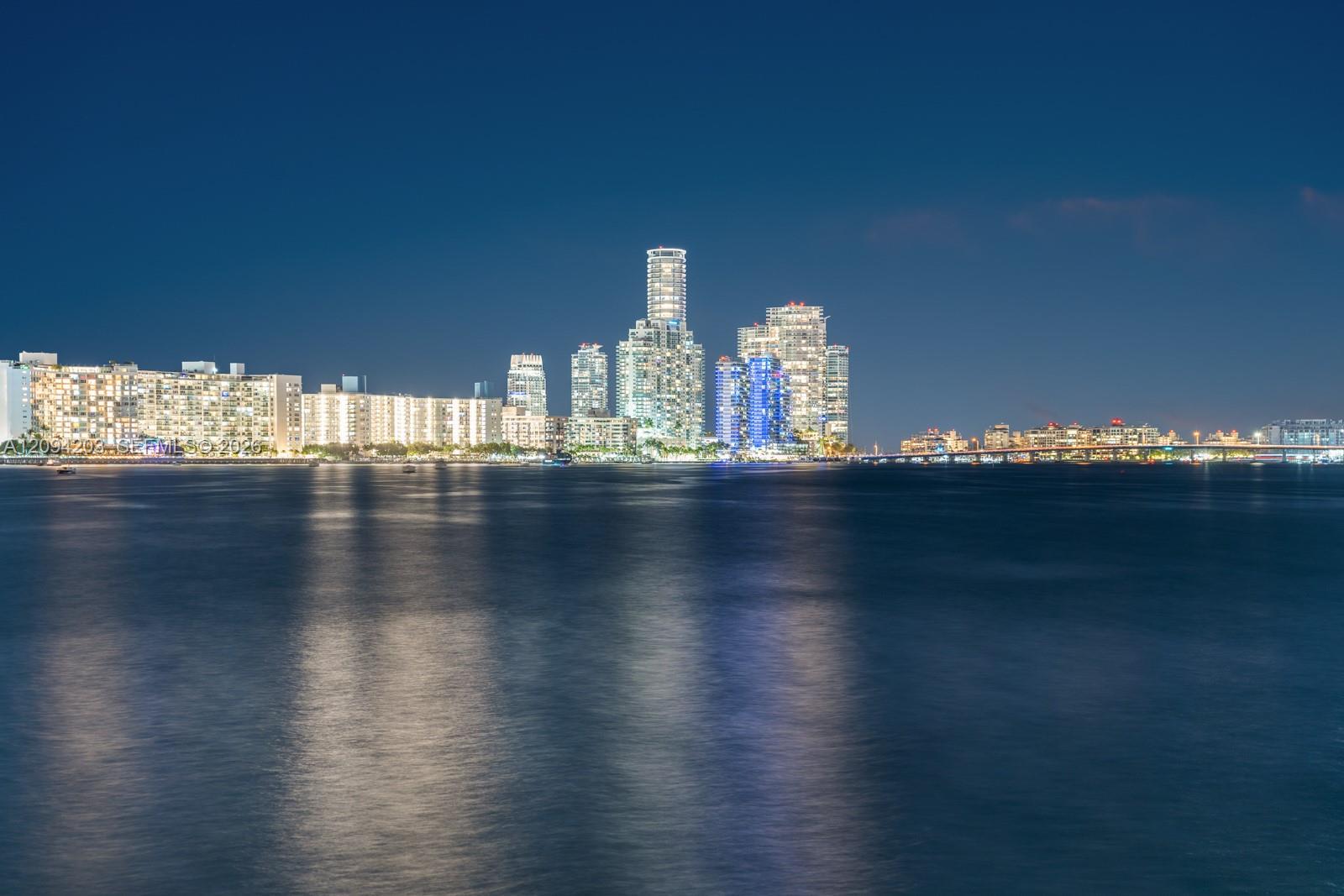 Miami skyline at night across the bay, with illuminated towers reflected on the water.