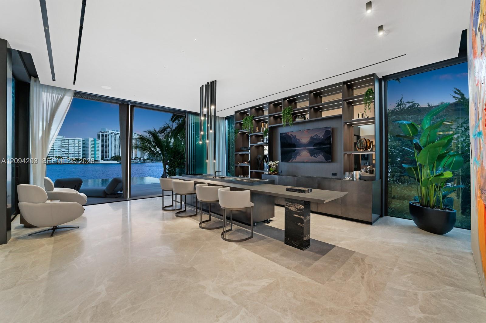 Waterfront great room with a stone island, built-in media shelving, lounge chairs, and floor-to-ceiling windows.