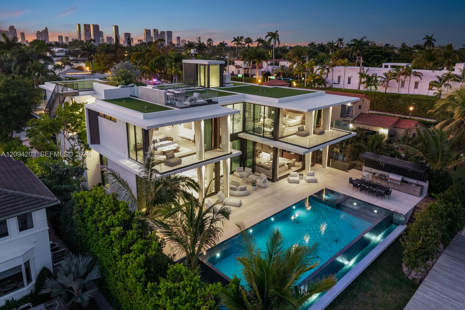 Modern glass residence with rooftop terrace and illuminated pool, framed by palms and the Miami skyline at dusk.