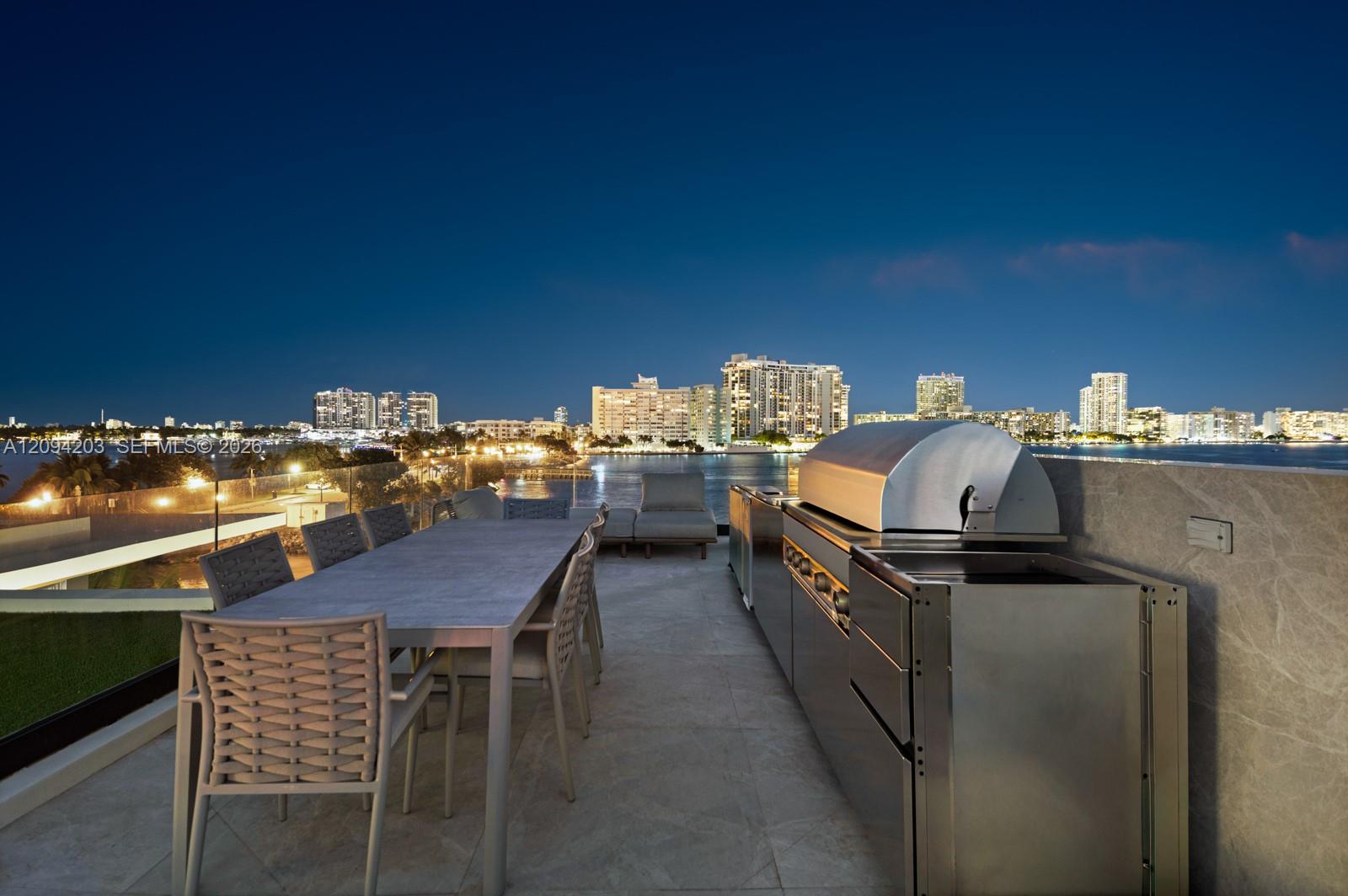 Rooftop terrace with an outdoor kitchen and dining area overlooking the water and illuminated skyline.