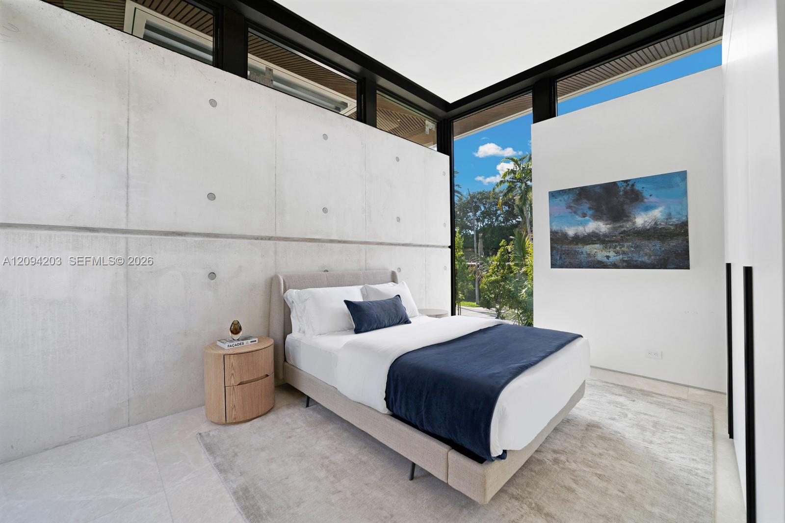 Modern bedroom with a concrete feature wall, navy bed linens, and floor-to-ceiling glass overlooking greenery.