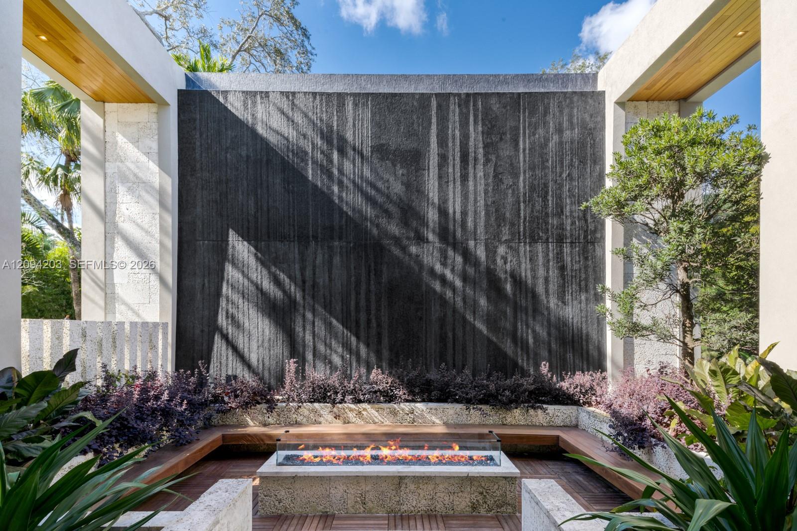 Modern courtyard with a tall dark water wall, tropical landscaping, and a long built-in fire feature.