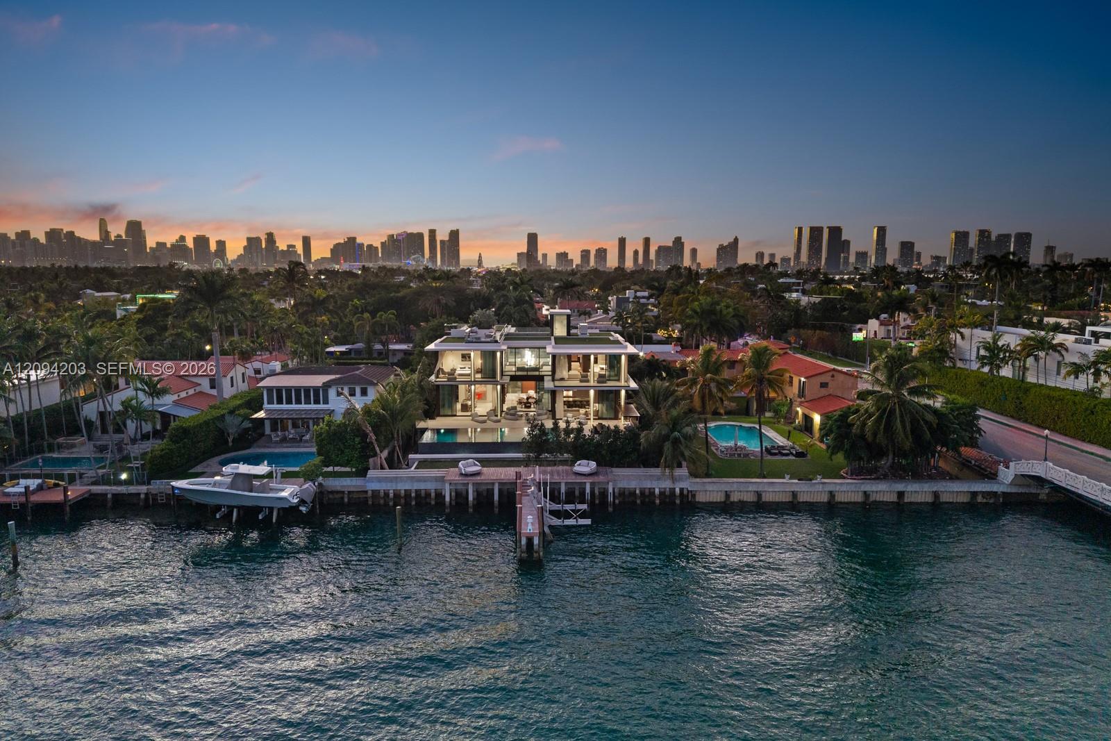 Aerial view of a modern waterfront home with a dock, pool, and Miami skyline at dusk.