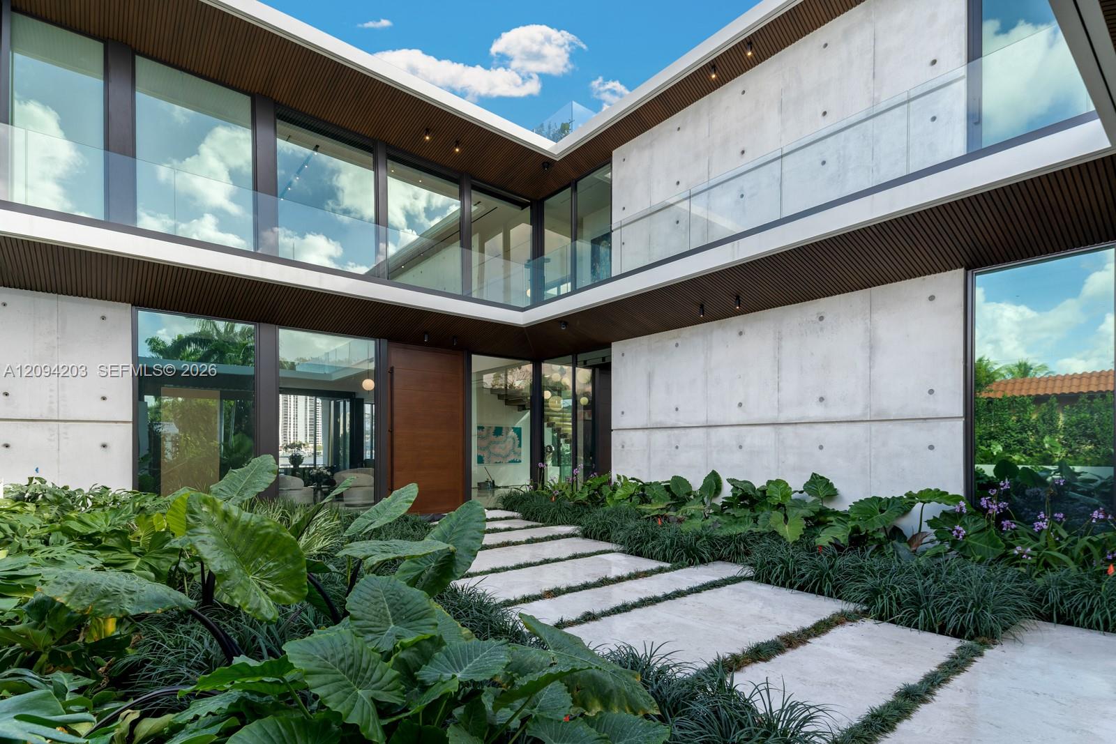 Modern concrete-and-glass home framed by tropical landscaping and a stepping-stone entrance path.