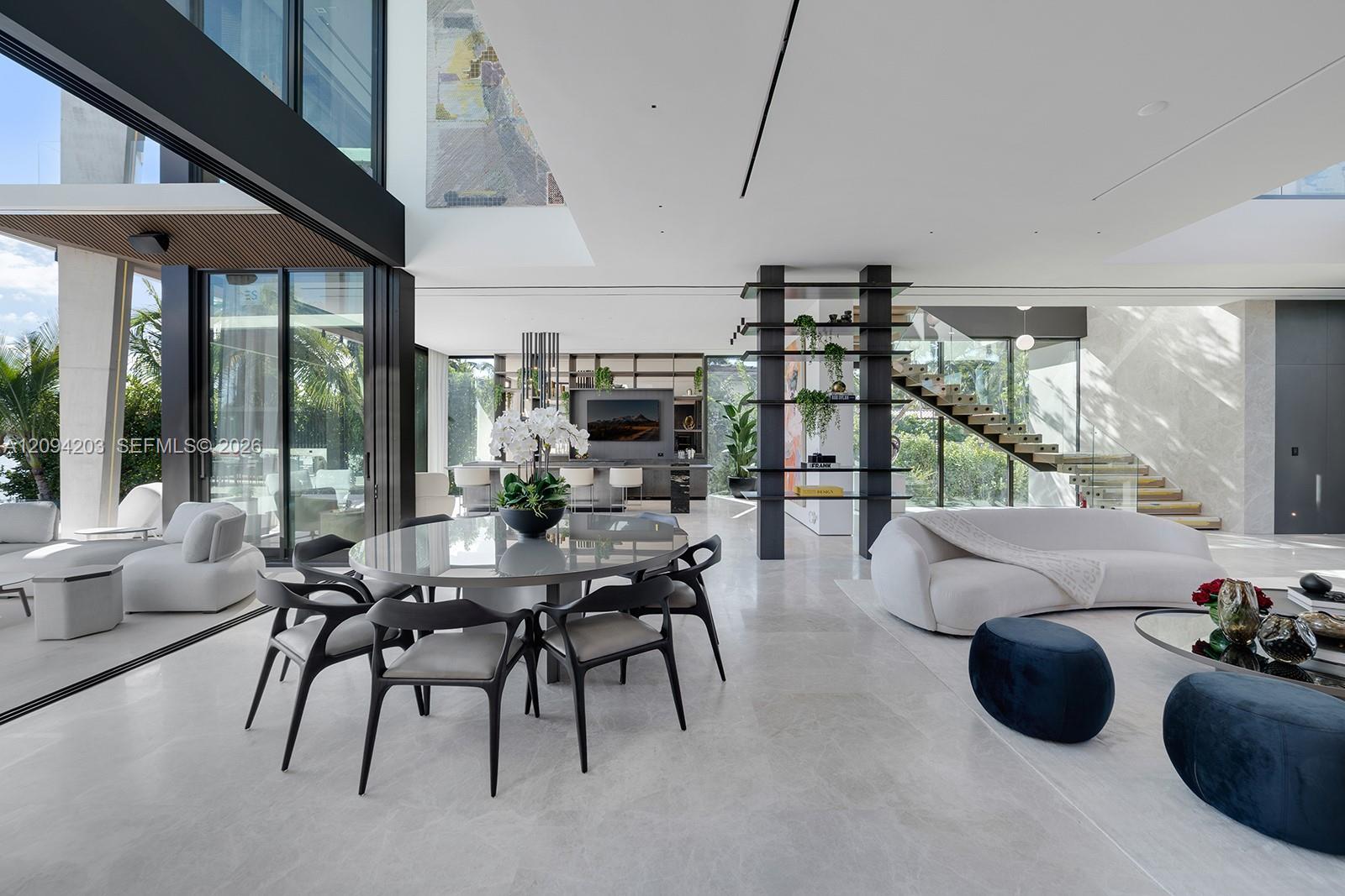 Modern living and dining room with white sofas, round dining table, open staircase, and glass walls.