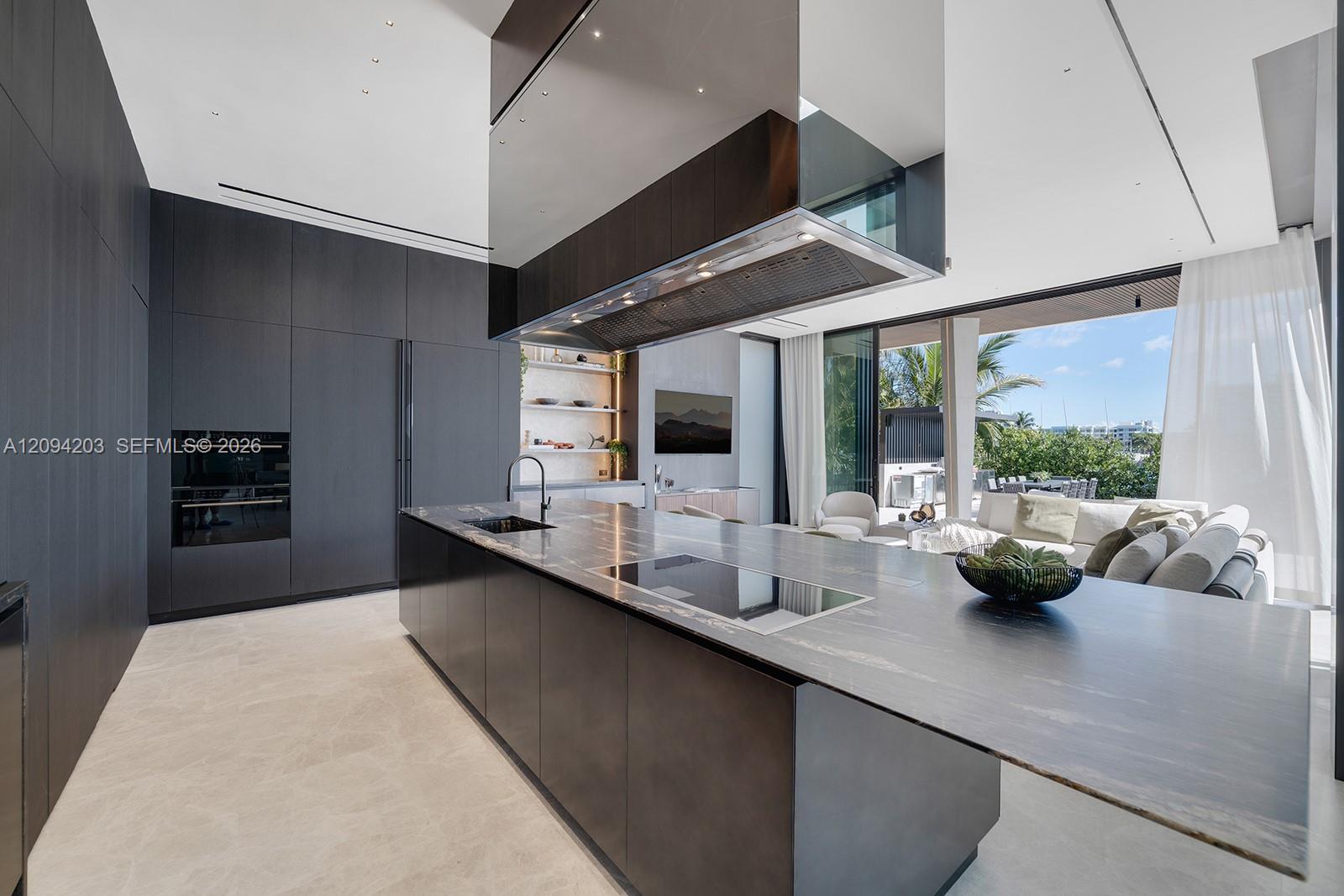 Modern kitchen with a long stone island, dark cabinetry, and windows opening to greenery.