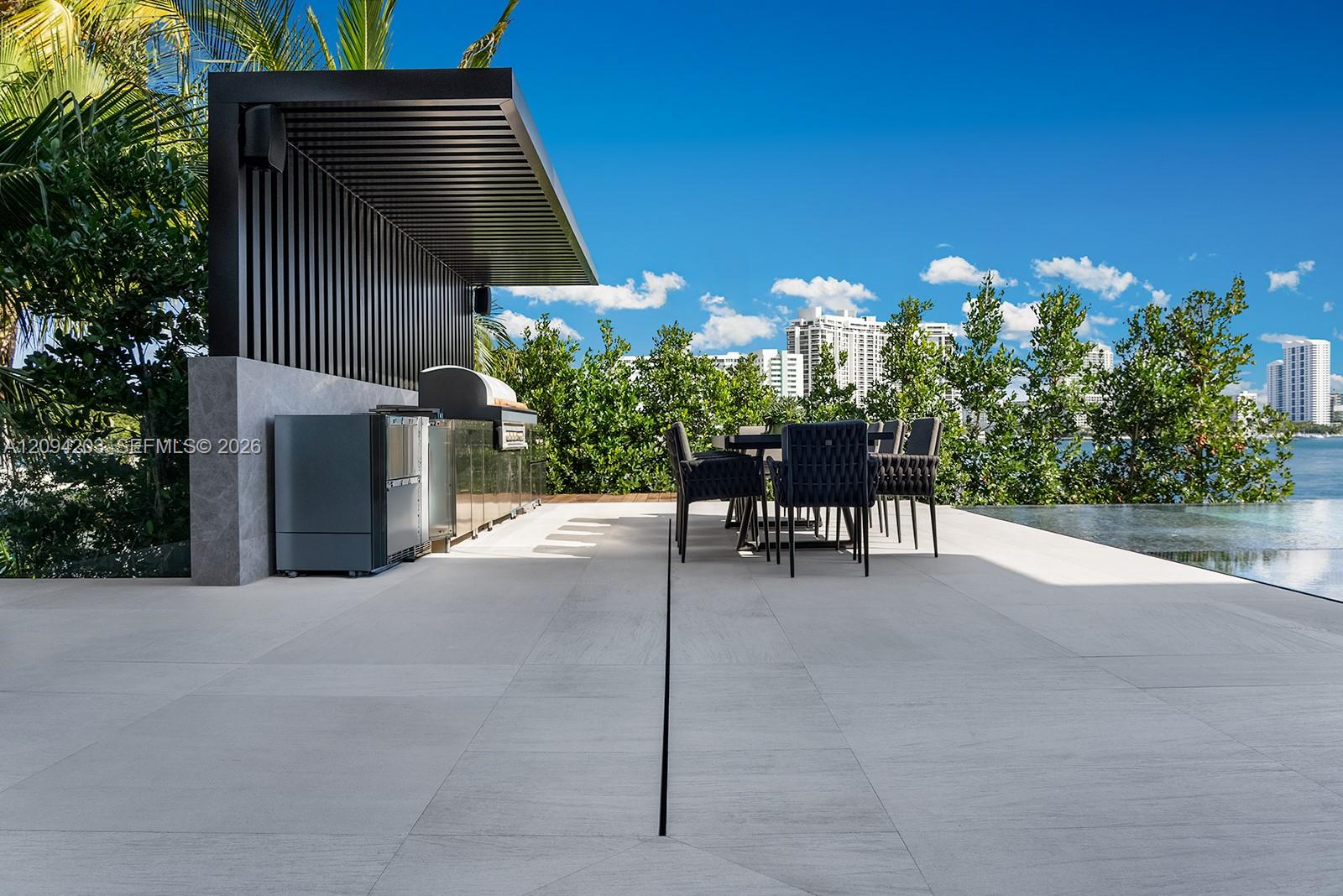 Waterfront patio with a modern outdoor kitchen, dining table, and infinity pool overlooking the Miami Beach skyline.