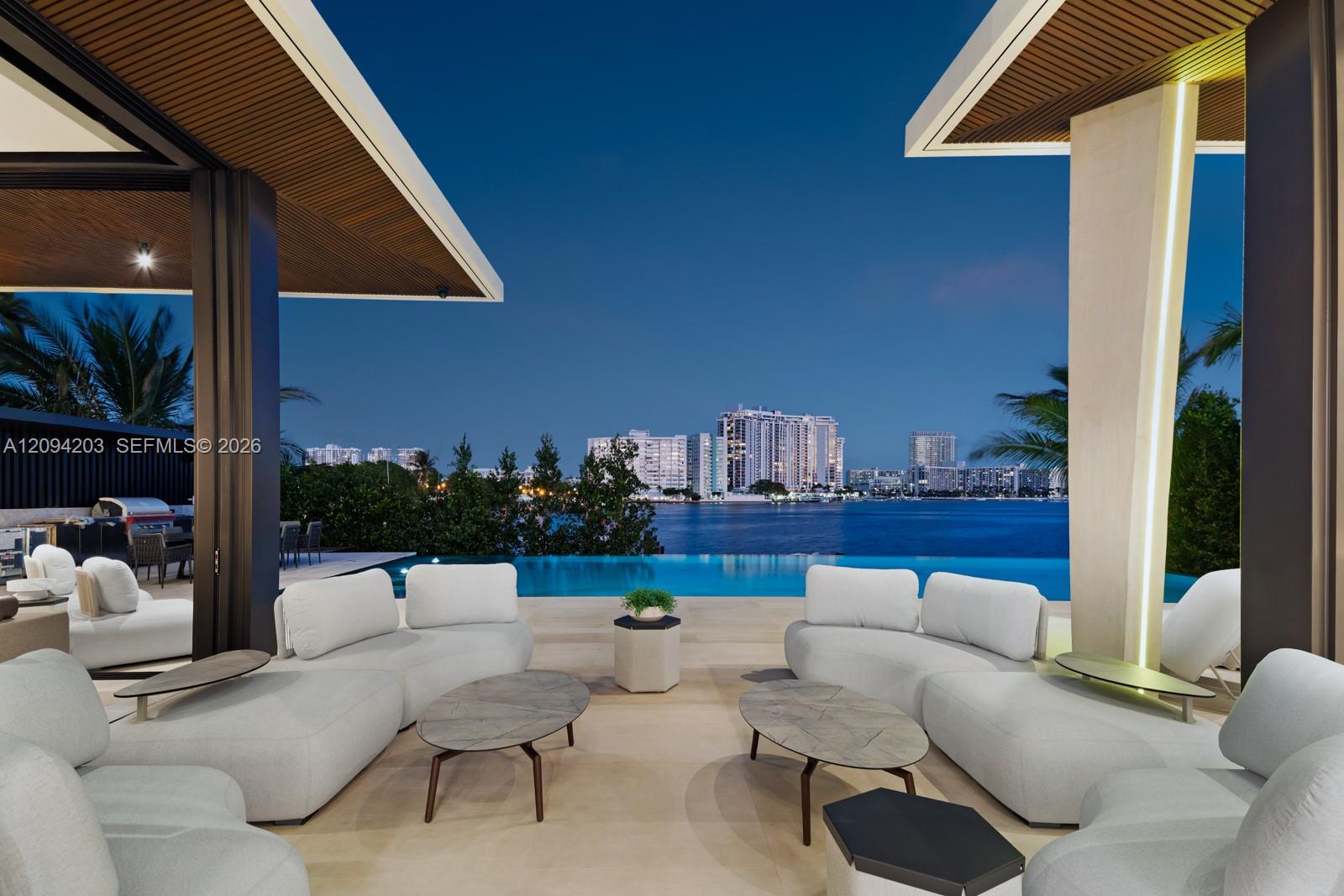Furnished patio with white sofas overlooking an infinity pool, bay, and Miami Beach skyline at dusk.