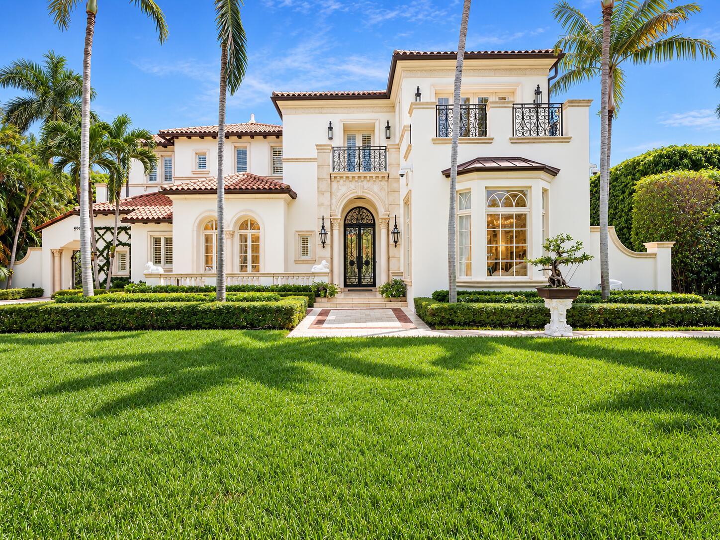 White two-story luxury home with terracotta roof, wrought-iron balconies, and palm-lined front lawn.