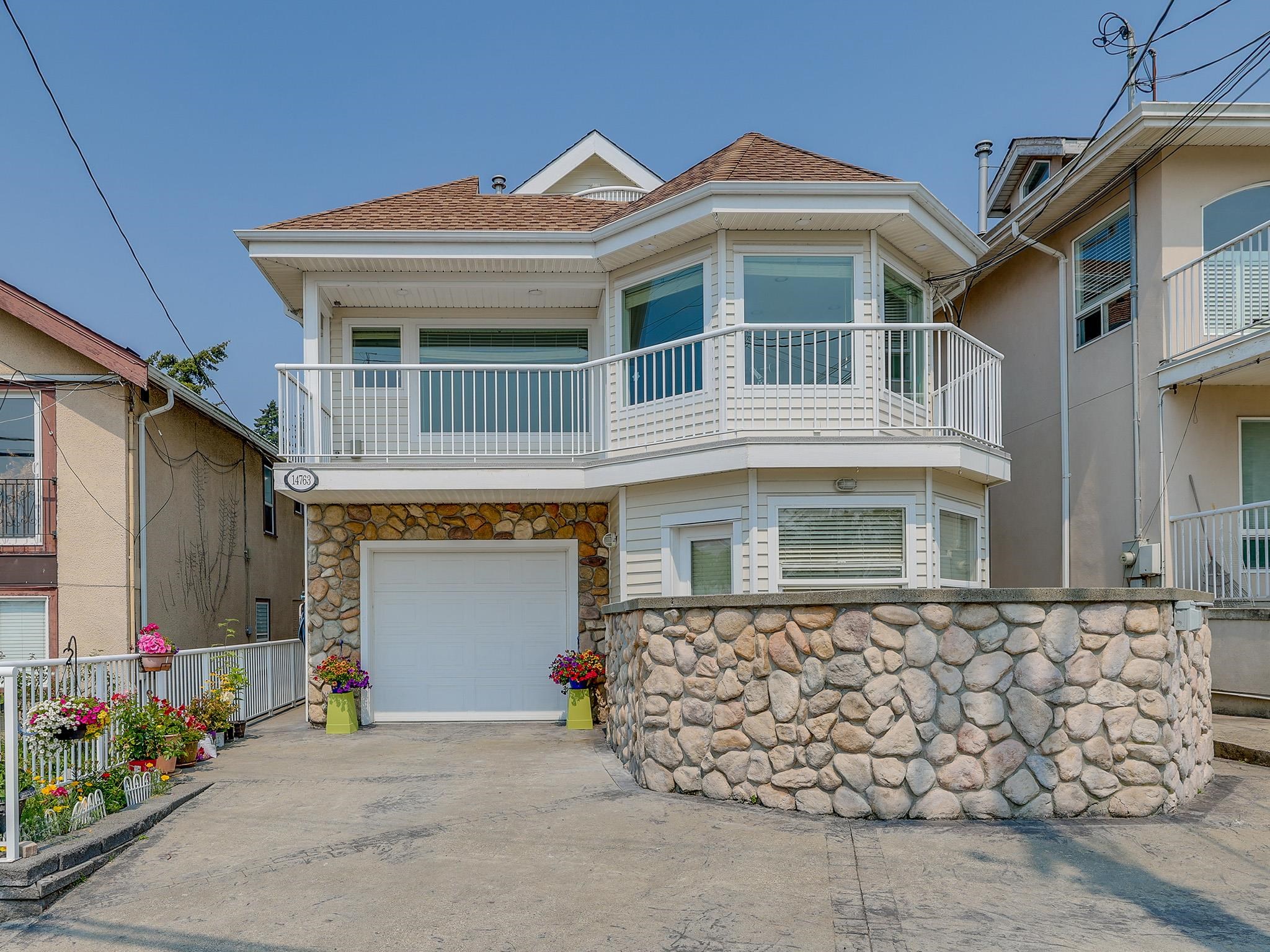 White two-story house with an upper balcony, stone garage facade, curved retaining wall, and flower planters.