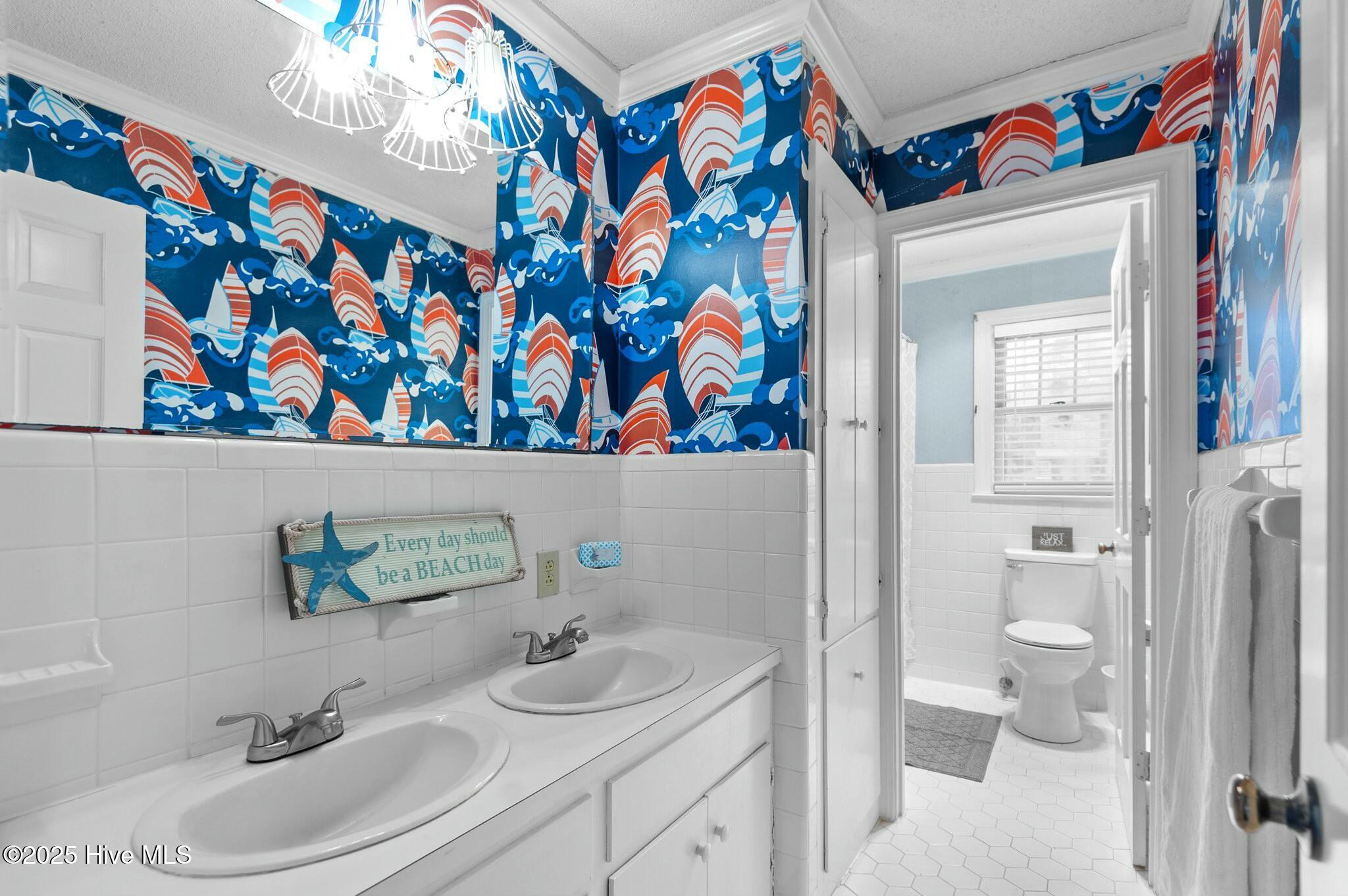 Bathroom with double sinks, sailboat wallpaper, and a doorway to a separate toilet area.