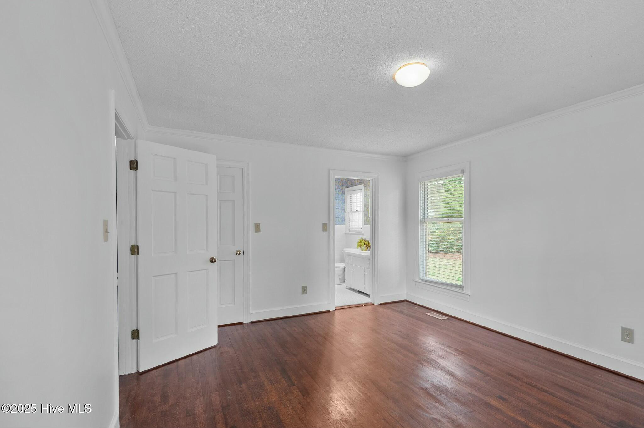 Empty room with dark hardwood floors, a window with blinds, and an open bathroom doorway.