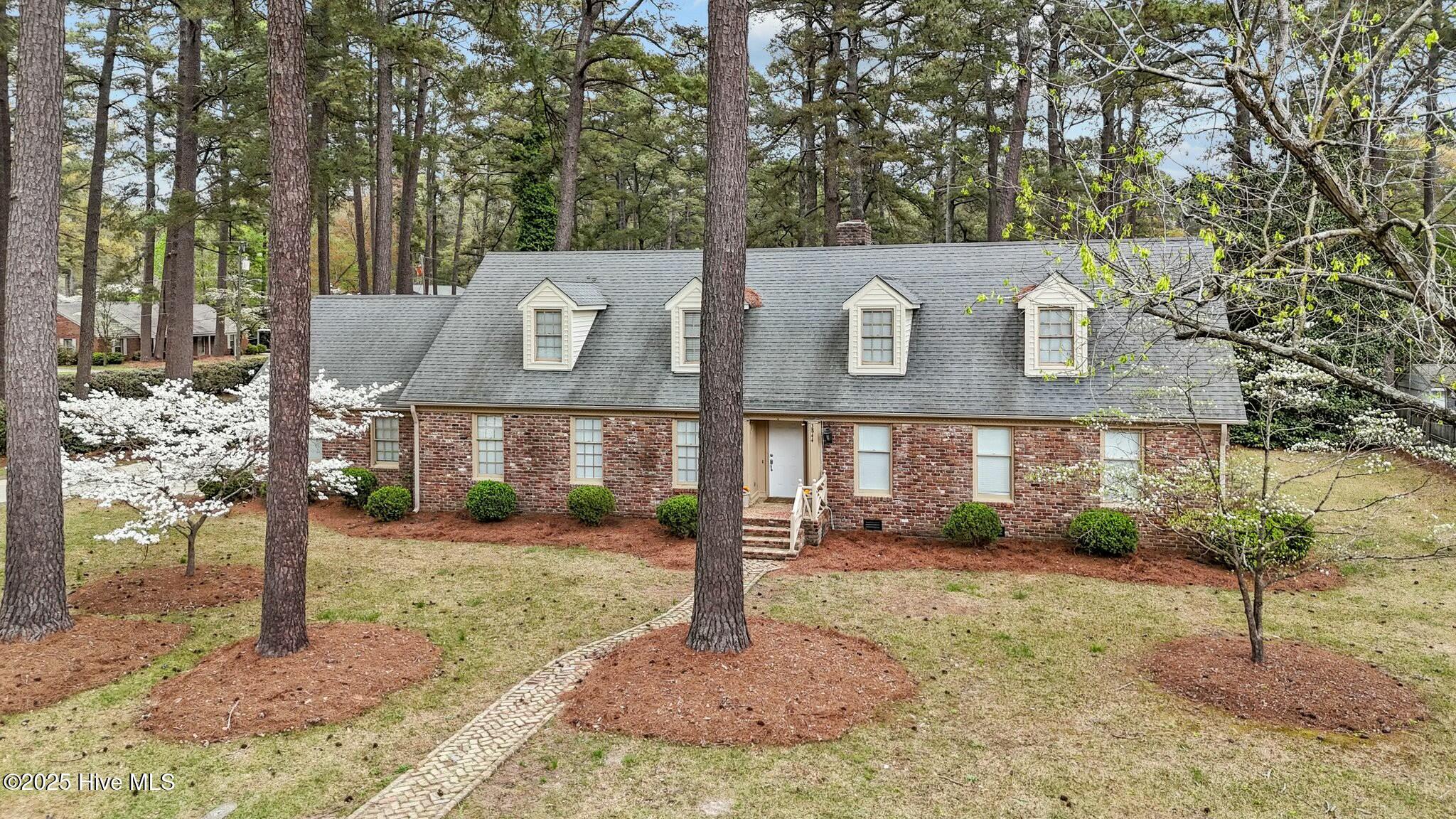 Brick house with three dormer windows, a curving stone path, and flowering trees among tall pines.