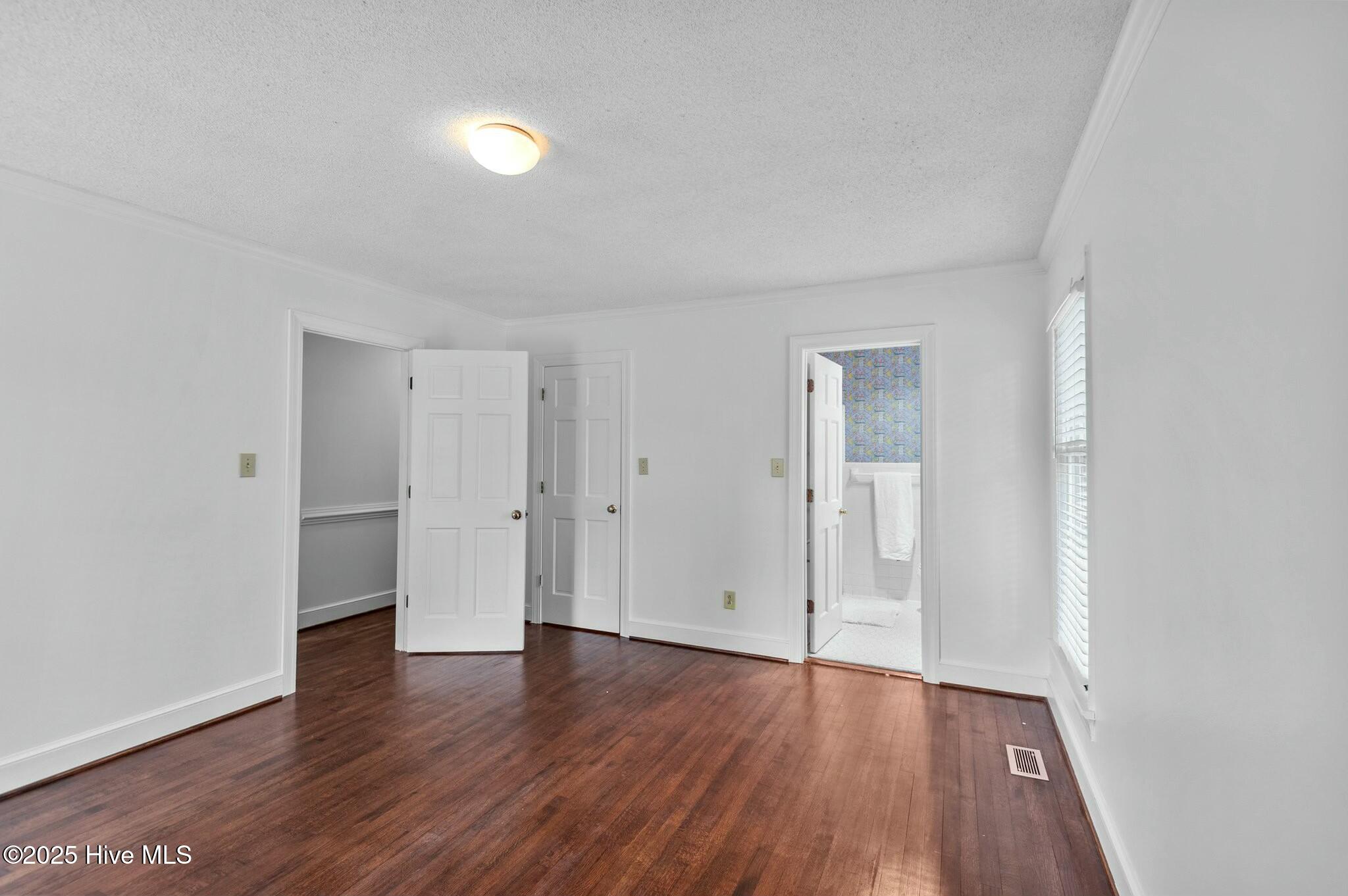 Empty room with dark hardwood floors, a window with blinds, and an open bathroom doorway.