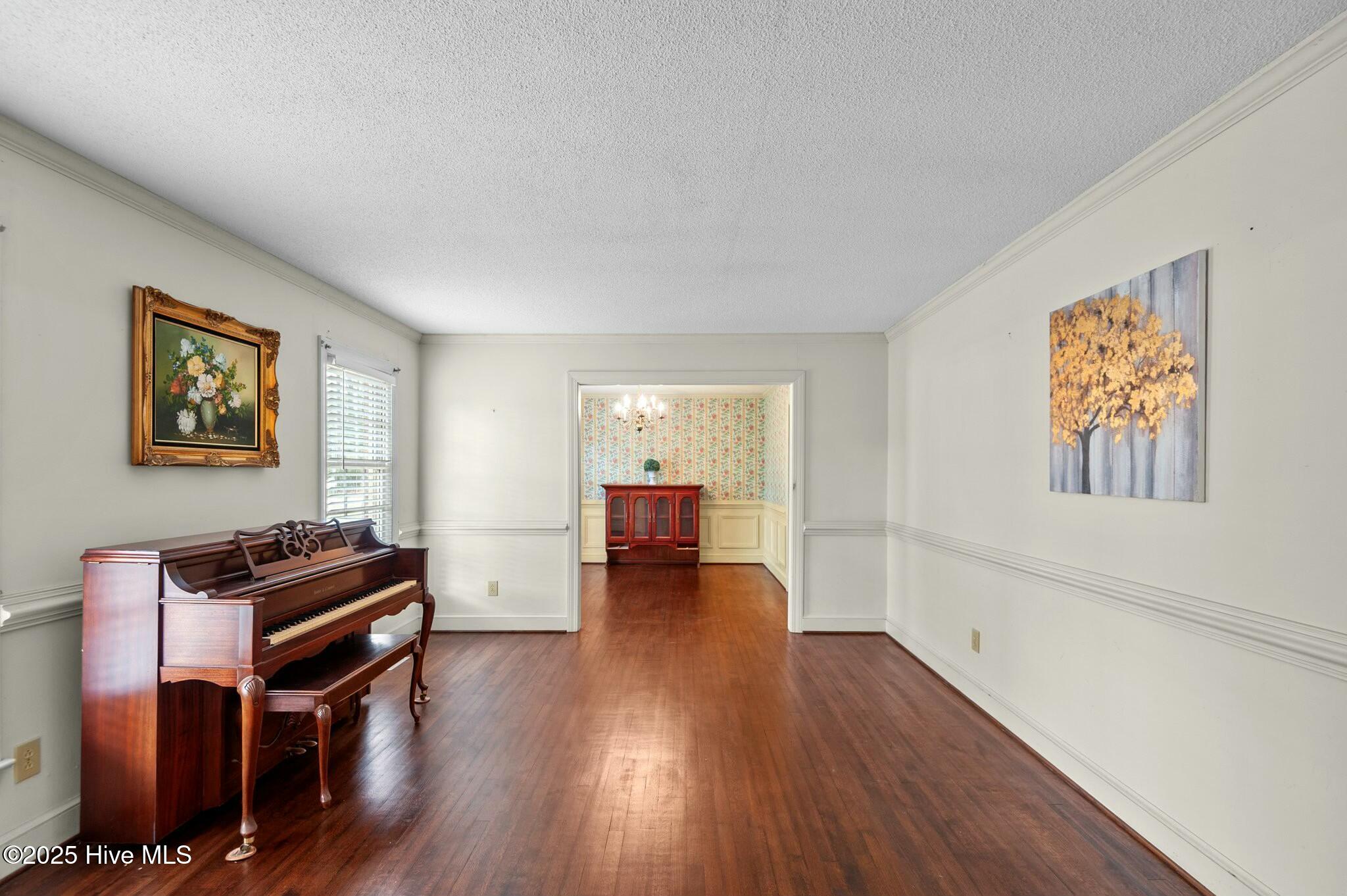 Furnished music room with a grand piano, polished wood floors, and a doorway to a floral-papered room.