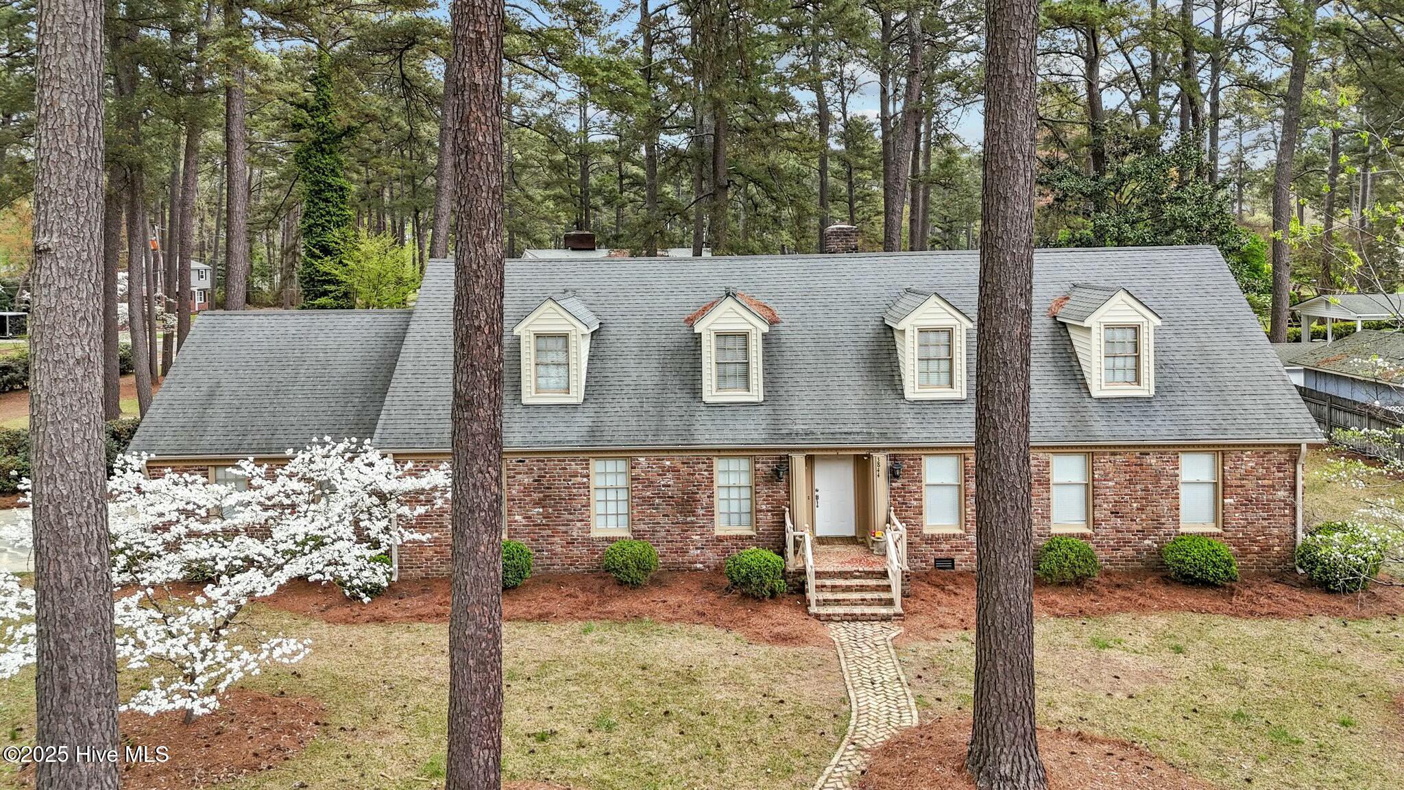 Brick house with four roof dormers, framed by tall pines and a flowering tree.
