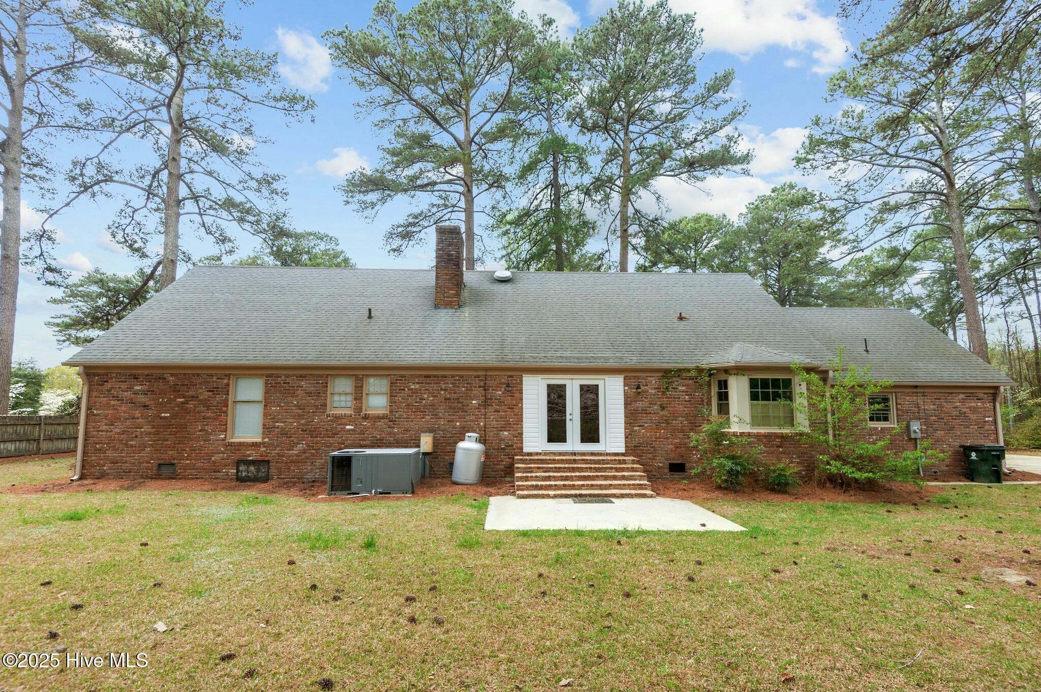 Red brick home’s rear exterior with white double doors, brick steps, and tall pine trees behind it.