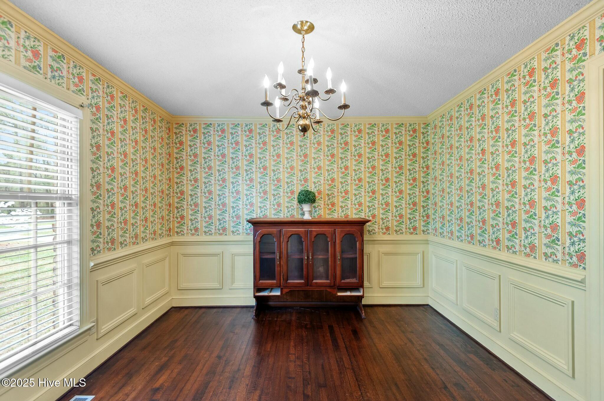 Dining room with floral wallpaper, pale wainscoting, dark hardwood floors, and a brass chandelier.