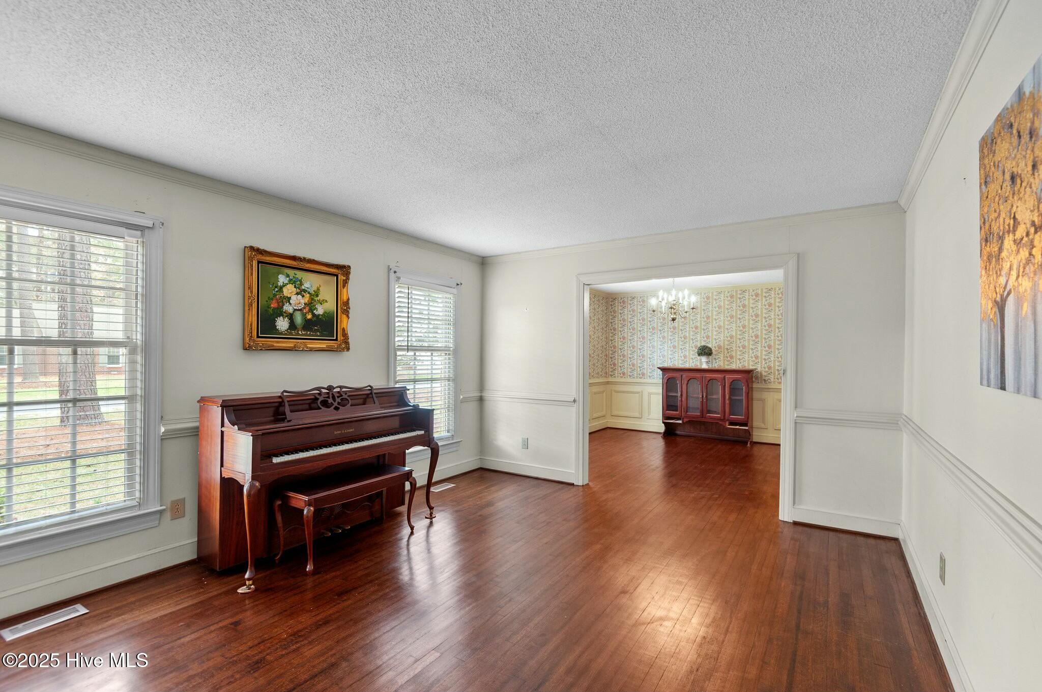 Front room with an upright piano, polished hardwood floors, and a wide doorway to a wallpapered adjoining room.