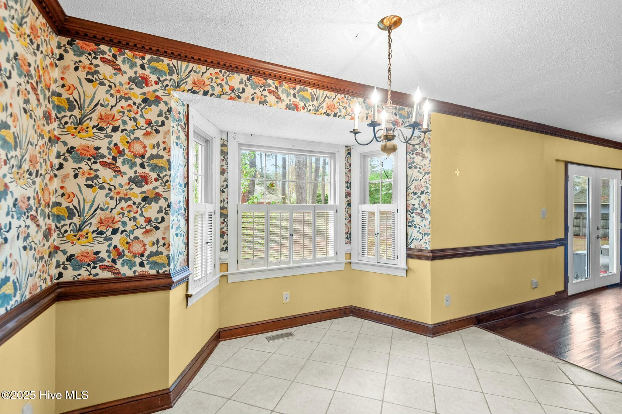 Dining area with a shuttered bay window, floral wallpaper, yellow walls, and a hanging chandelier.