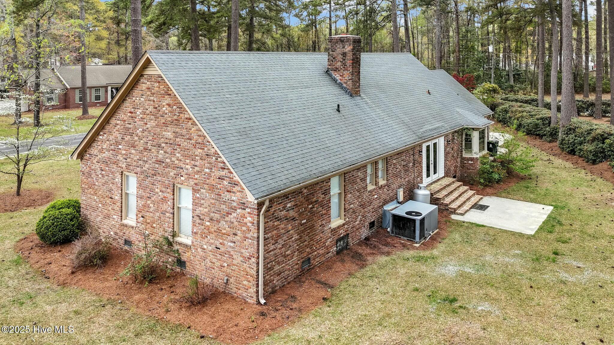 Rear and side of a brick house with a broad gray roof and chimney, surrounded by trees.