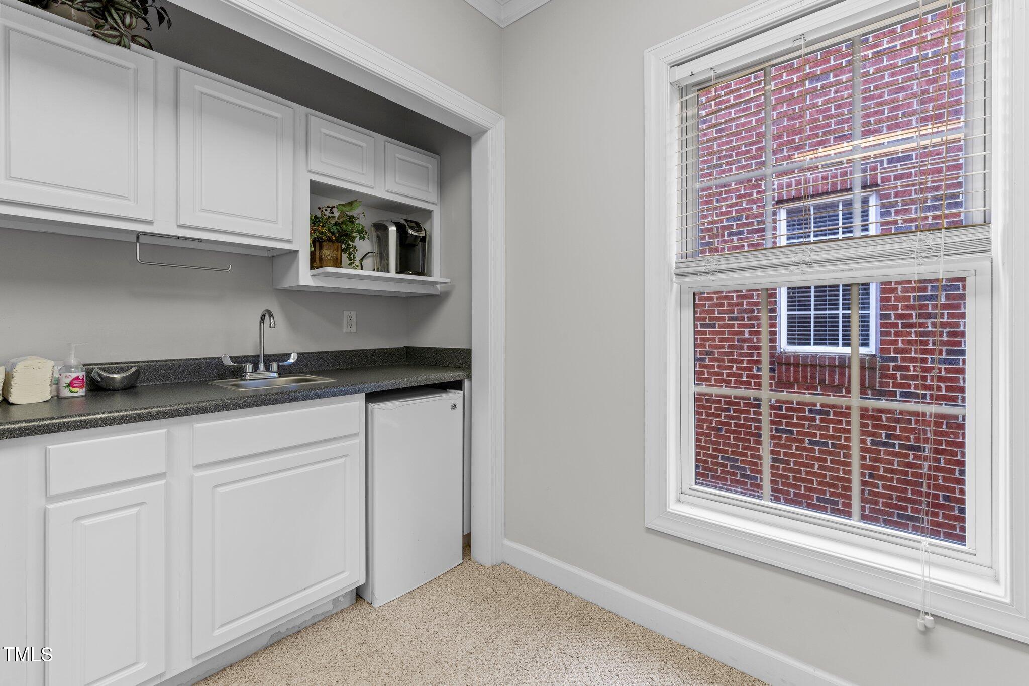 Compact kitchenette with white cabinets, dark counters, a small refrigerator, and a window facing a brick wall.