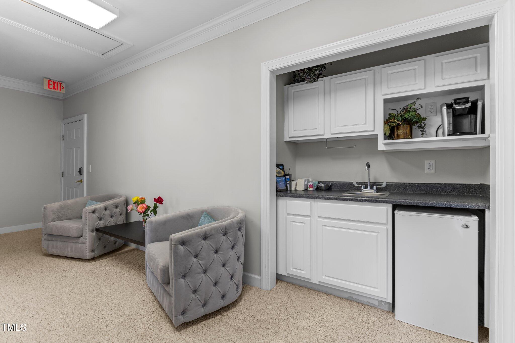 Furnished lounge with gray tufted armchairs beside a kitchenette with white cabinets and a compact refrigerator.