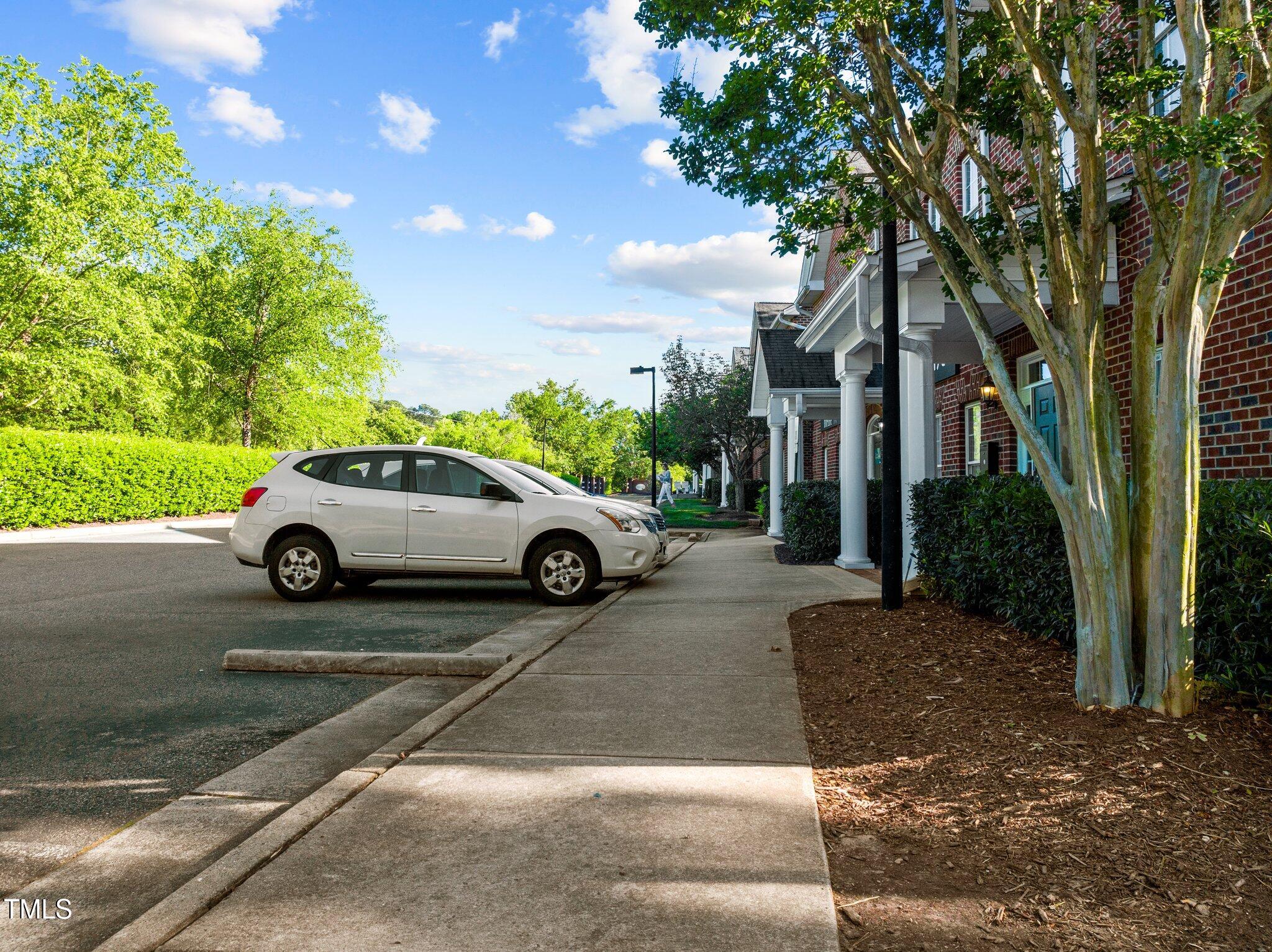 Brick condominium entrance beside a sidewalk, landscaped trees, and a parked white SUV.