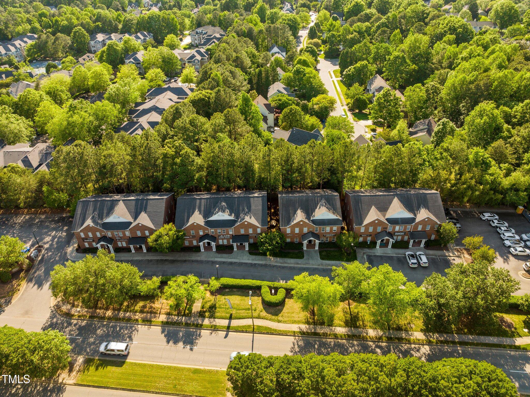 Aerial view of brick condominium buildings beside a tree-lined road and wooded neighborhood.