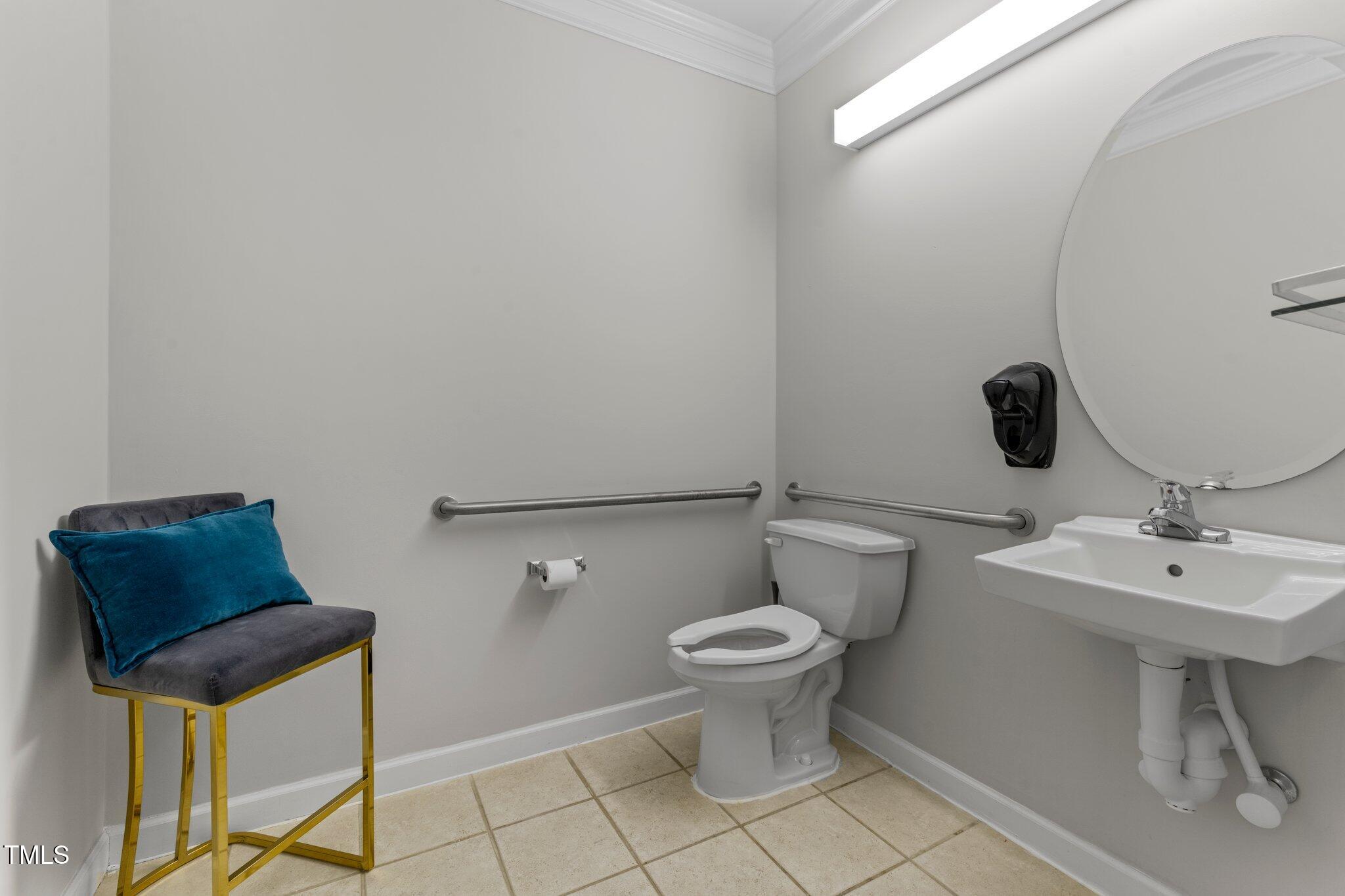 Bathroom with grab bars, a round mirror, wall-mounted sink, and a blue-cushioned chair.