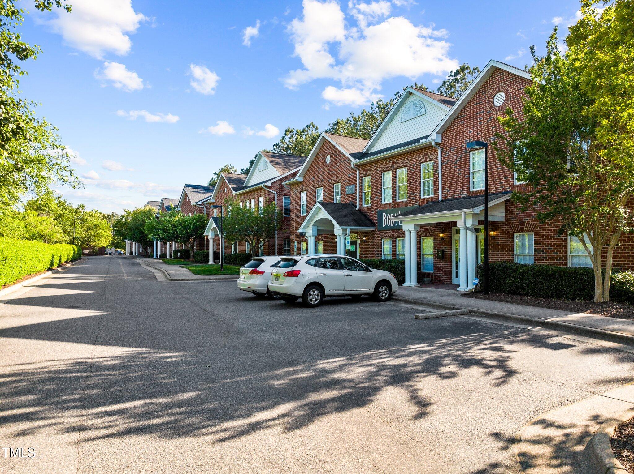 Brick multi-unit building with white gables, covered entrances, and parked cars along the driveway.