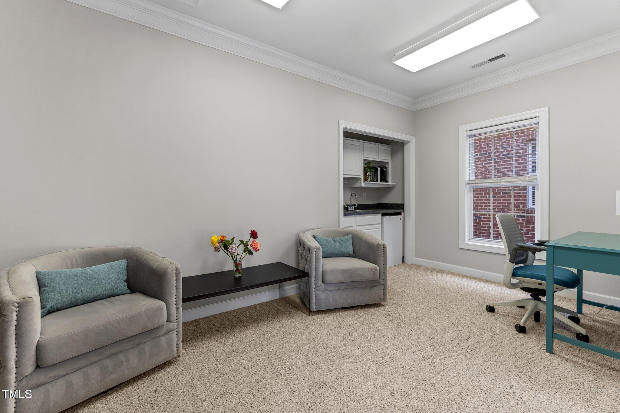 Furnished office with gray armchairs, teal desk, kitchenette, and a window facing a brick wall.