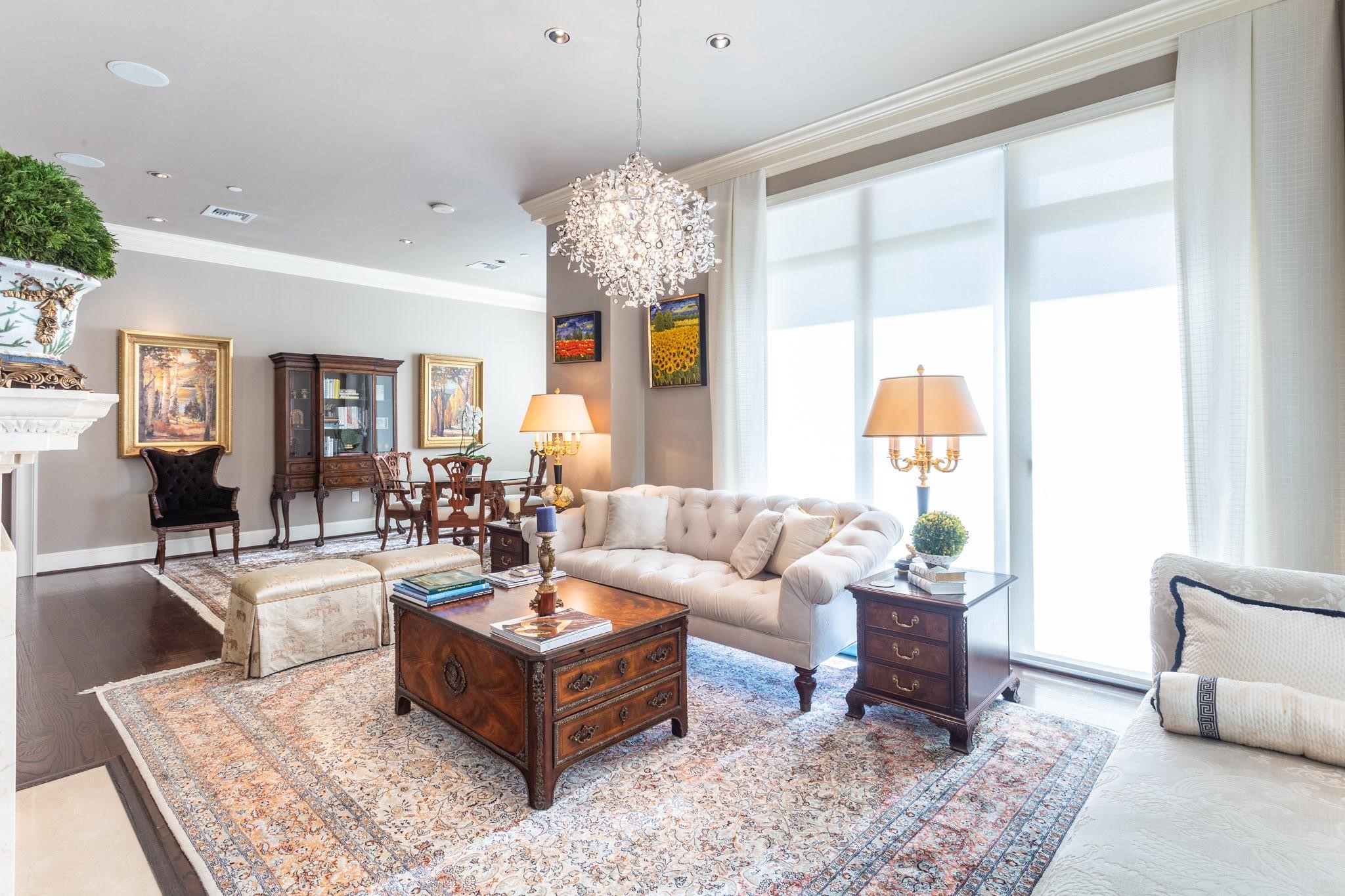 Living room with a cream tufted sofa, ornate wood furniture, crystal chandelier, and tall shaded windows.