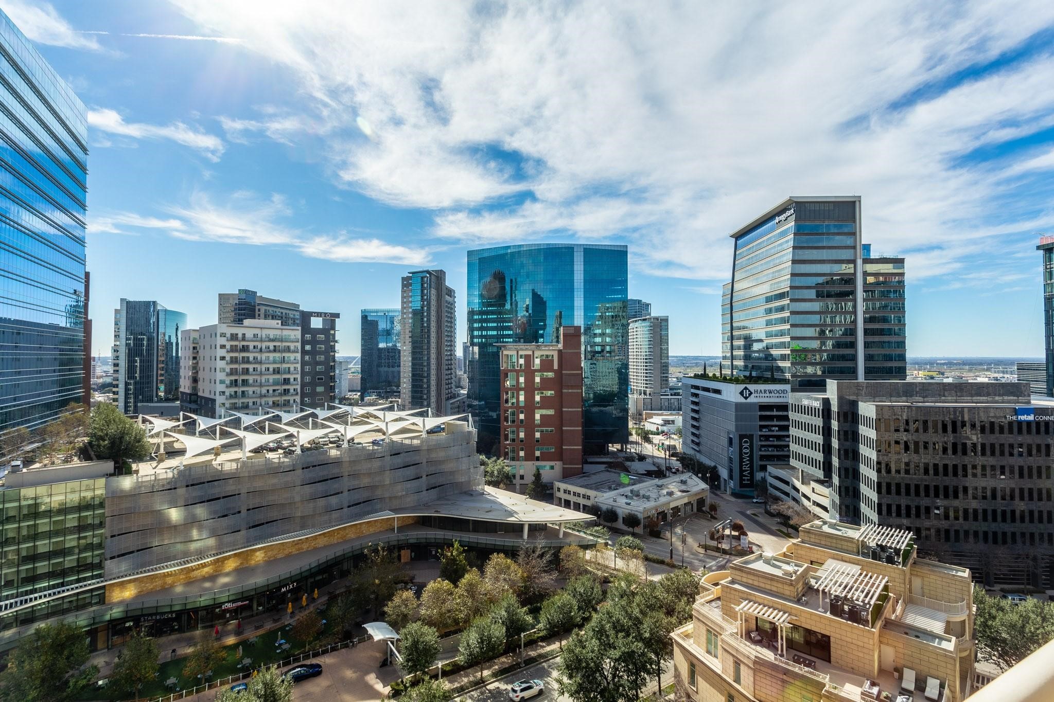 Downtown Dallas skyline with glass towers, rooftop terraces, and a tree-lined street.