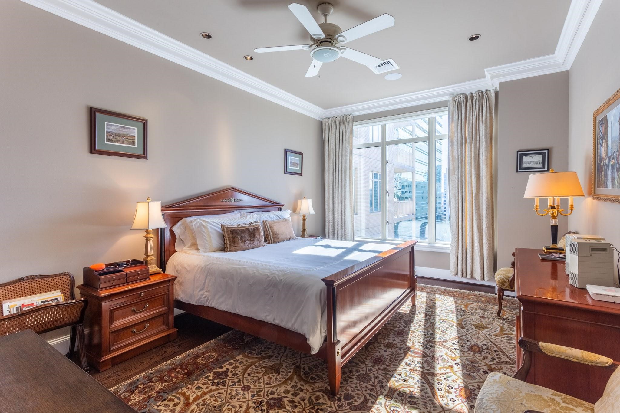 Furnished bedroom with a dark wood bed, patterned rug, and large windows overlooking nearby buildings.